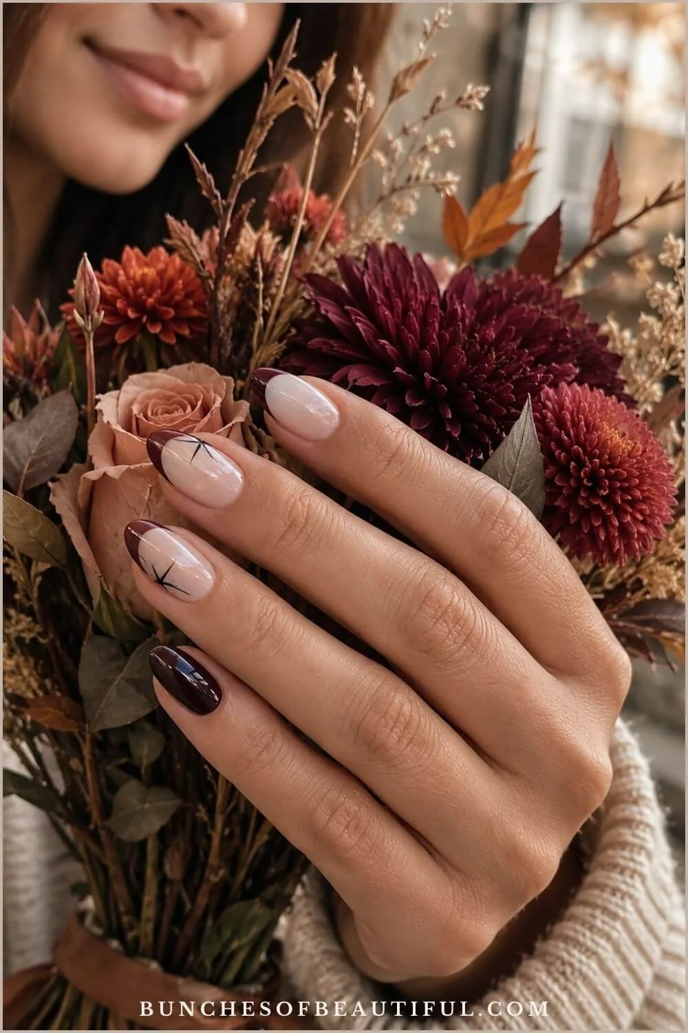 Nude almond nails with burgundy French tips and fine black spike accents posed over an autumn flower bouquet.