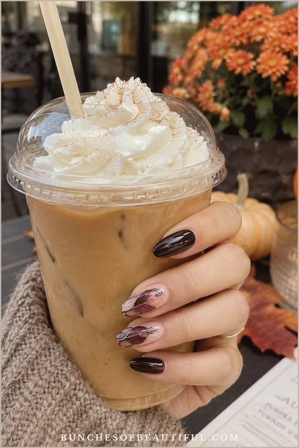 Burgundy and nude almond nails with stained-glass-style gothic accents while holding an iced coffee near fall flowers and pumpkins.