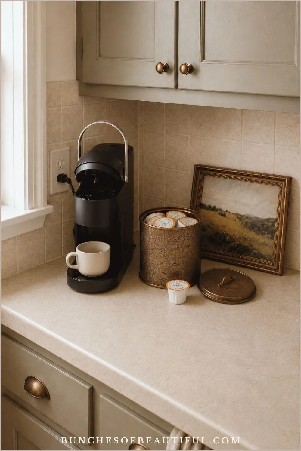 Compact pod coffee corner with a narrow black brewer, cream mug, open metal canister filled with coffee pods, and vintage landscape art.
