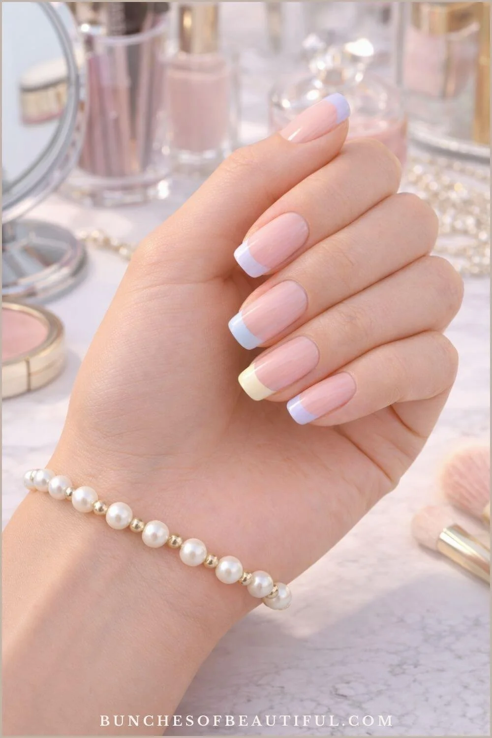 Spring pastel French tip nails with lavender, yellow, and blue tips on a natural pink base photographed with pearls and vanity items.