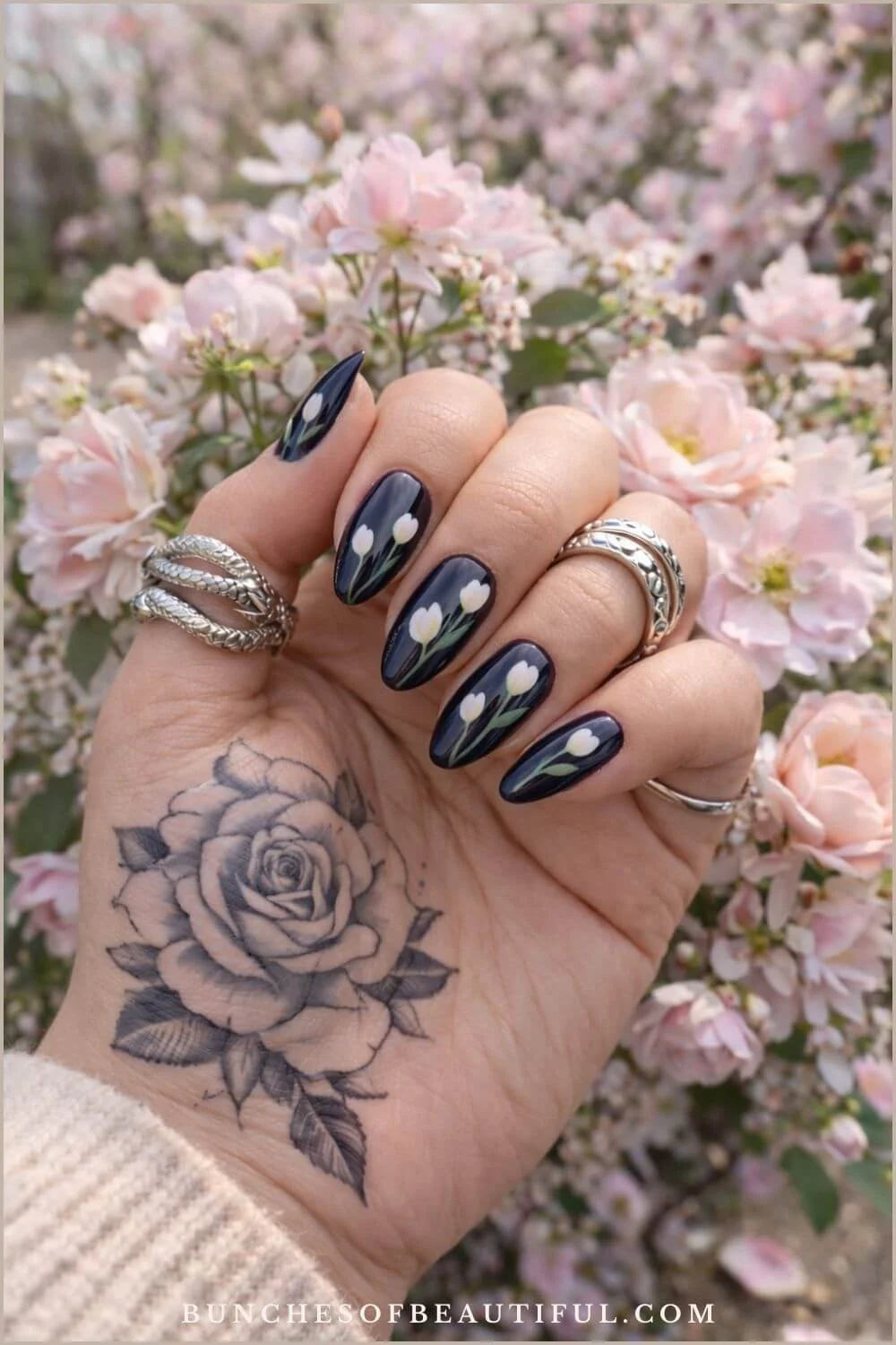 Dark navy spring nails with white tulip nail art on long almond nails photographed in front of blooming pink spring flowers.