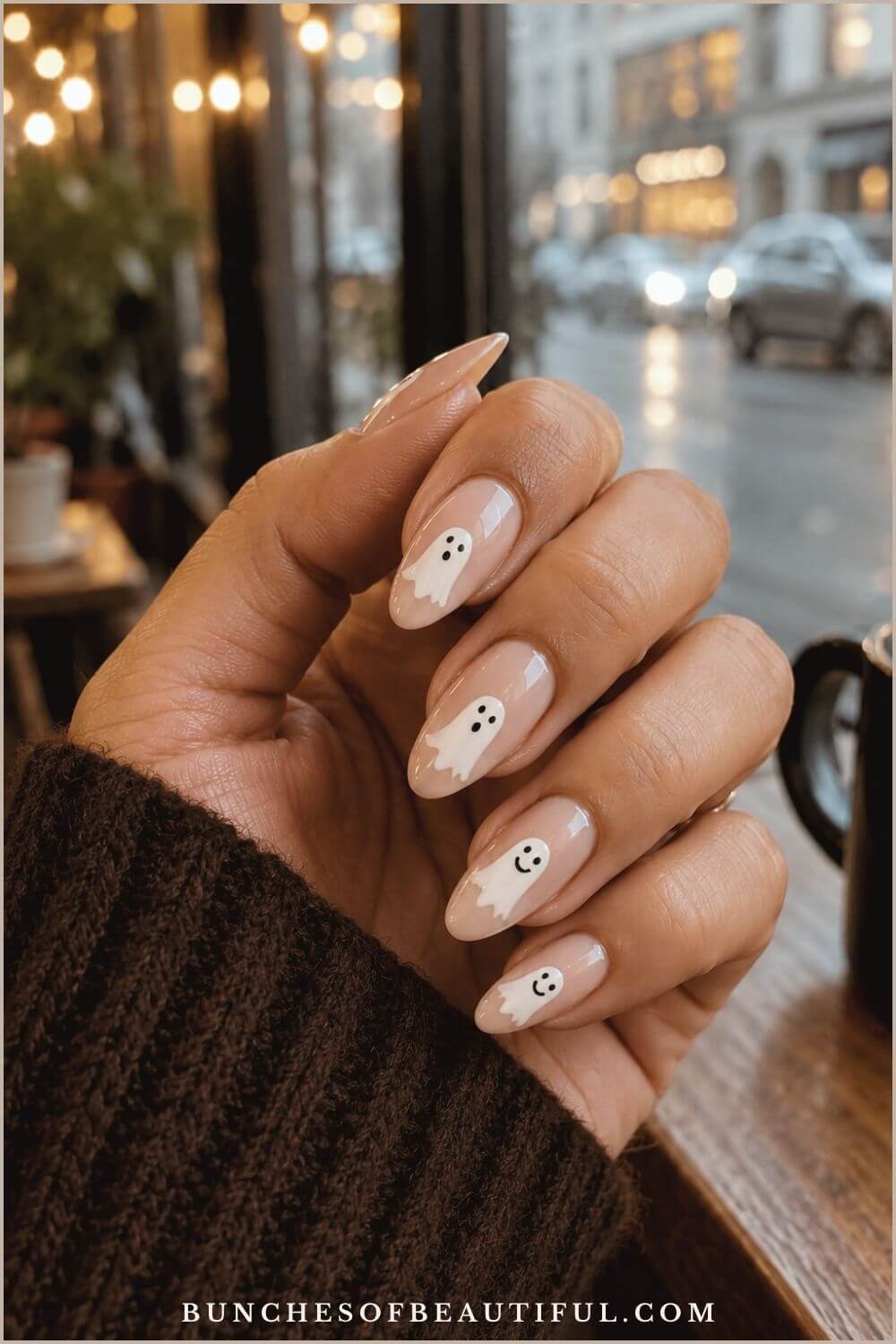 Nude almond nails decorated with playful white ghosts in a cozy café beside a rainy window.