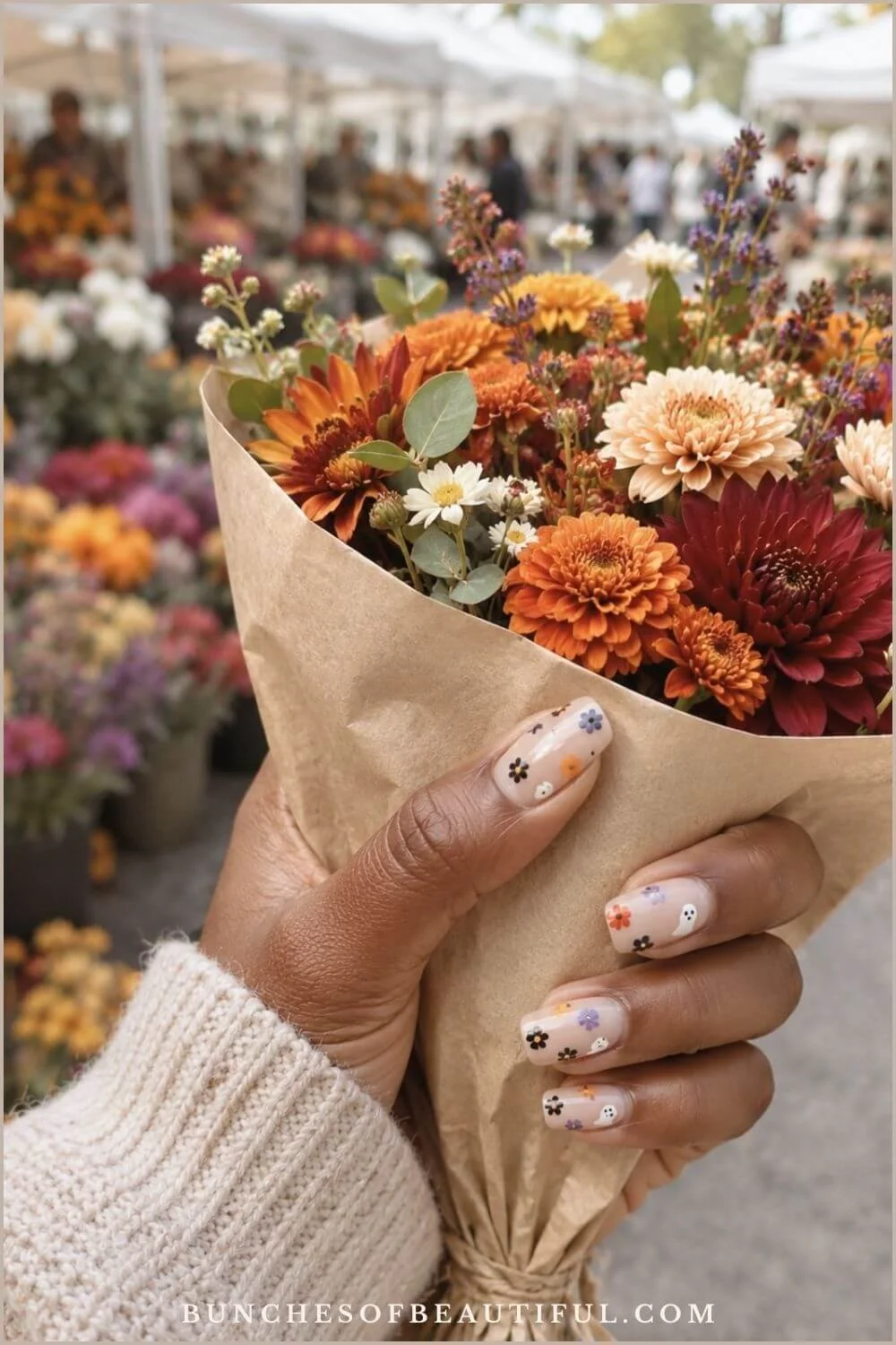 Short nude nails with tiny autumn flowers and white ghosts while holding a bouquet at an outdoor flower market.