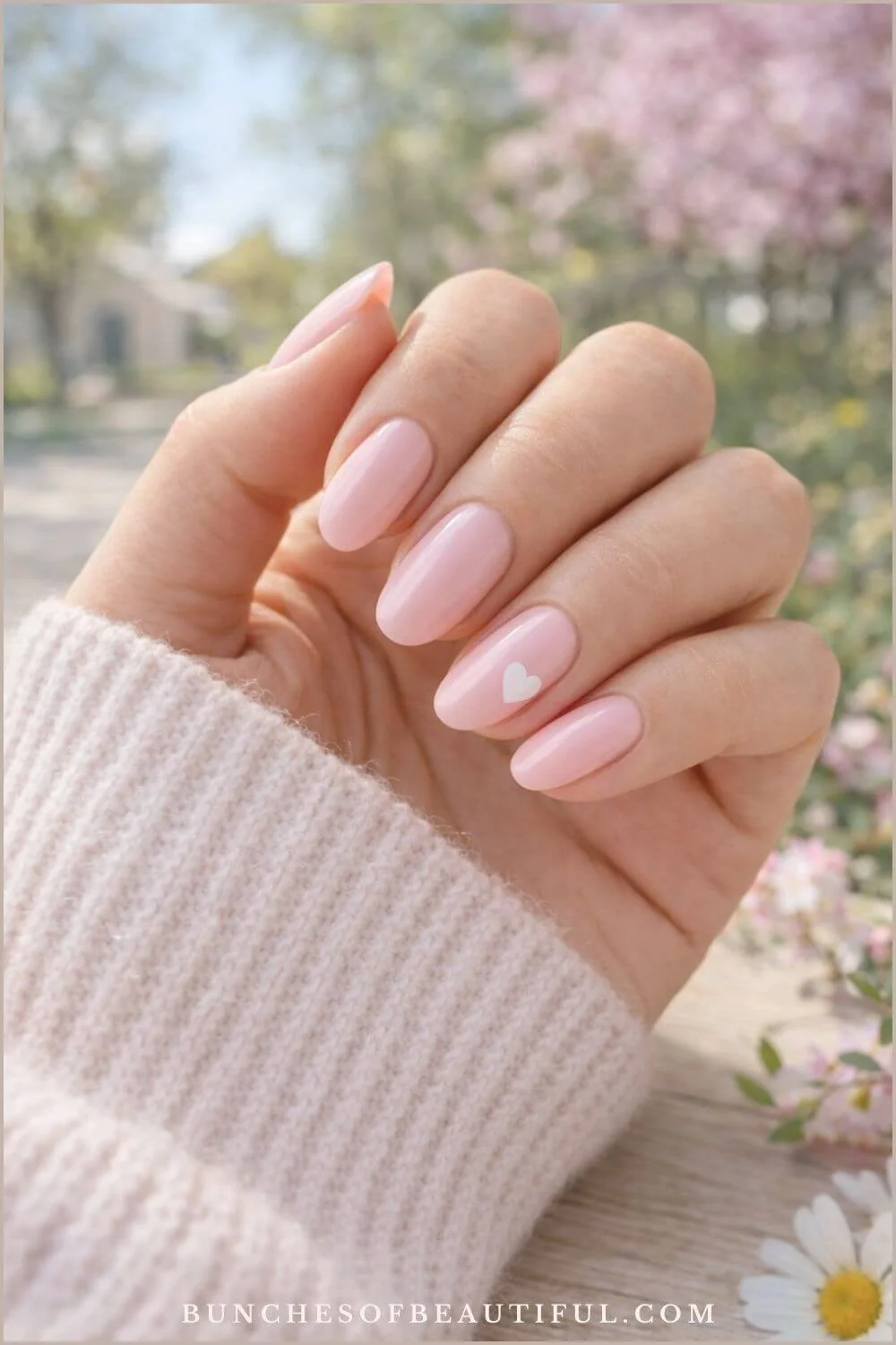 Pastel pink spring nails with a small white heart accent on almond shaped glossy nails photographed with soft spring flowers in the background.