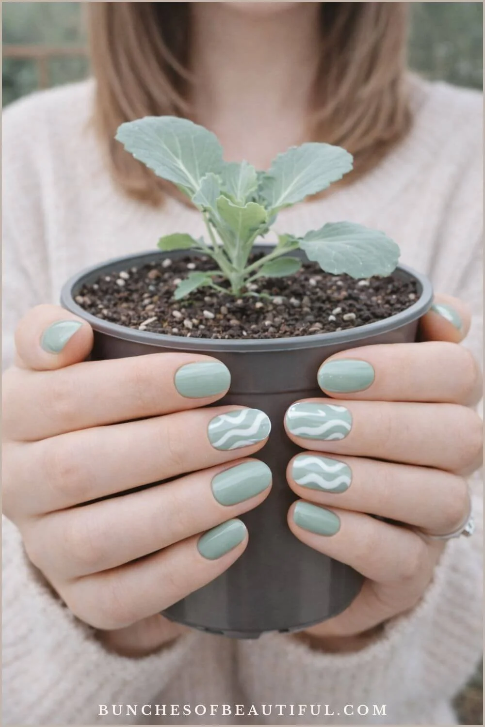 Short sage green spring nails with subtle white wavy accents while holding a small plant in a black pot.