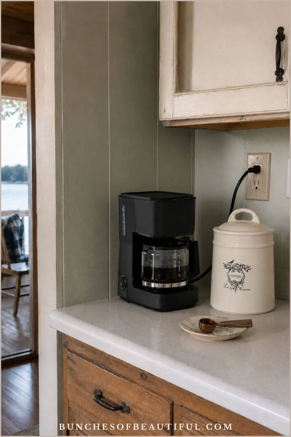 Small rustic counter with a black drip coffee maker, white ceramic coffee canister, scoop, muted green wall, and natural wood cabinetry.