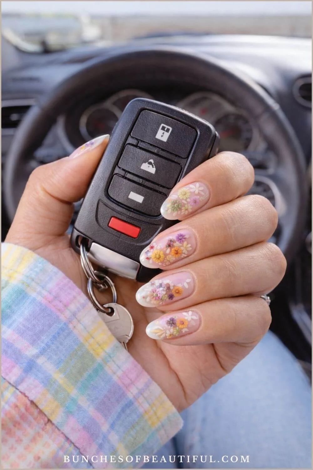 Sheer spring nails with delicate pressed flower nail art on almond shaped nails, photographed inside a car in natural daylight.