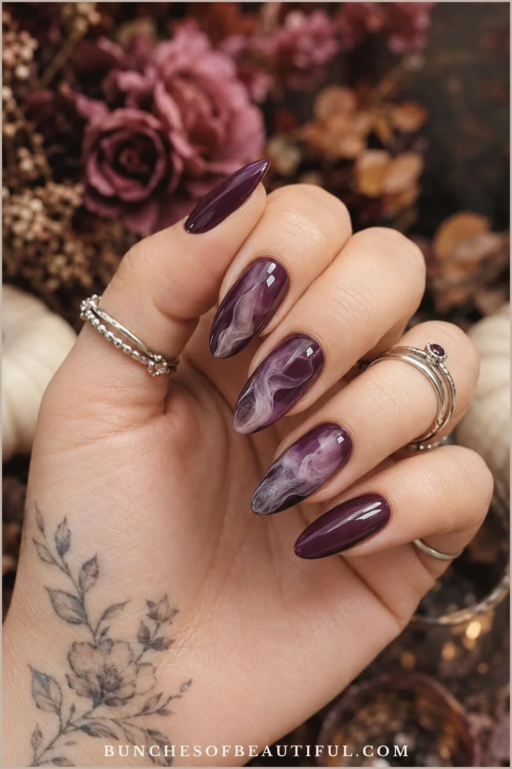 Deep plum almond nails with wispy white smoke accents, silver rings, and dried burgundy flowers in the background.