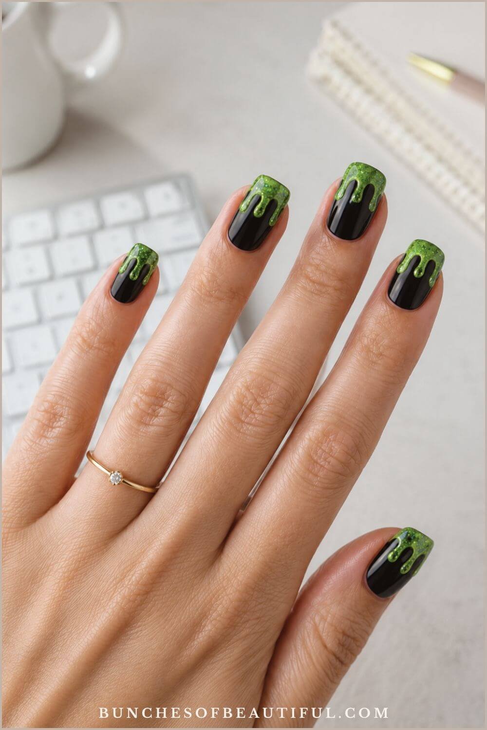 Glossy black square nails with glittery green slime dripping from the tips beside a white computer keyboard.