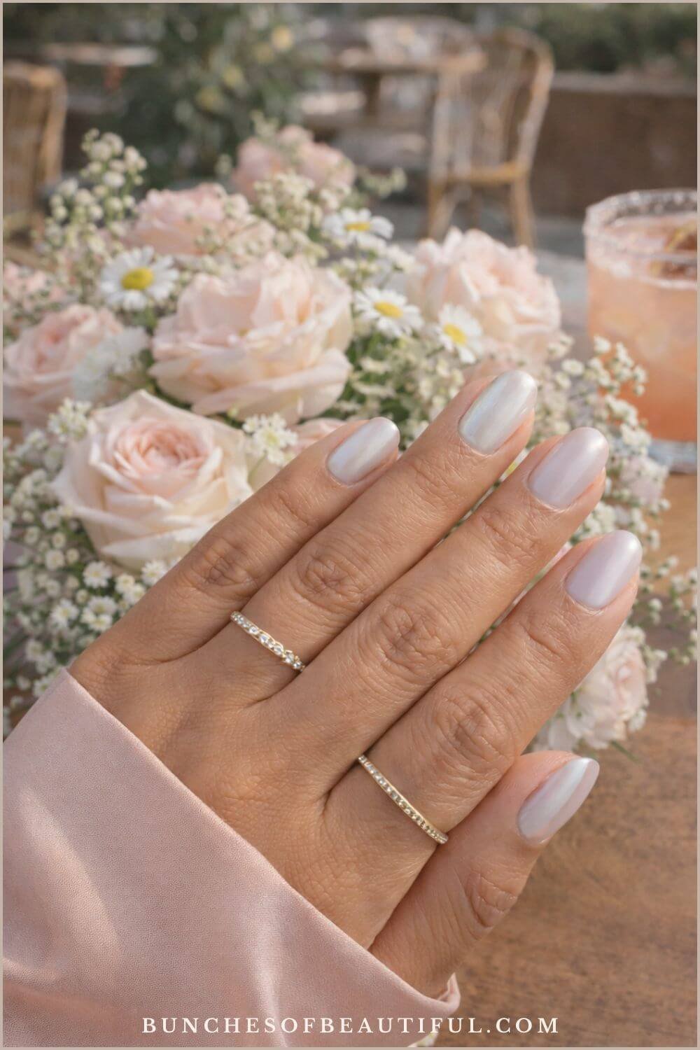 Soft pearl spring chrome nails on short rounded nails photographed outdoors with blush roses and daisies in the background.