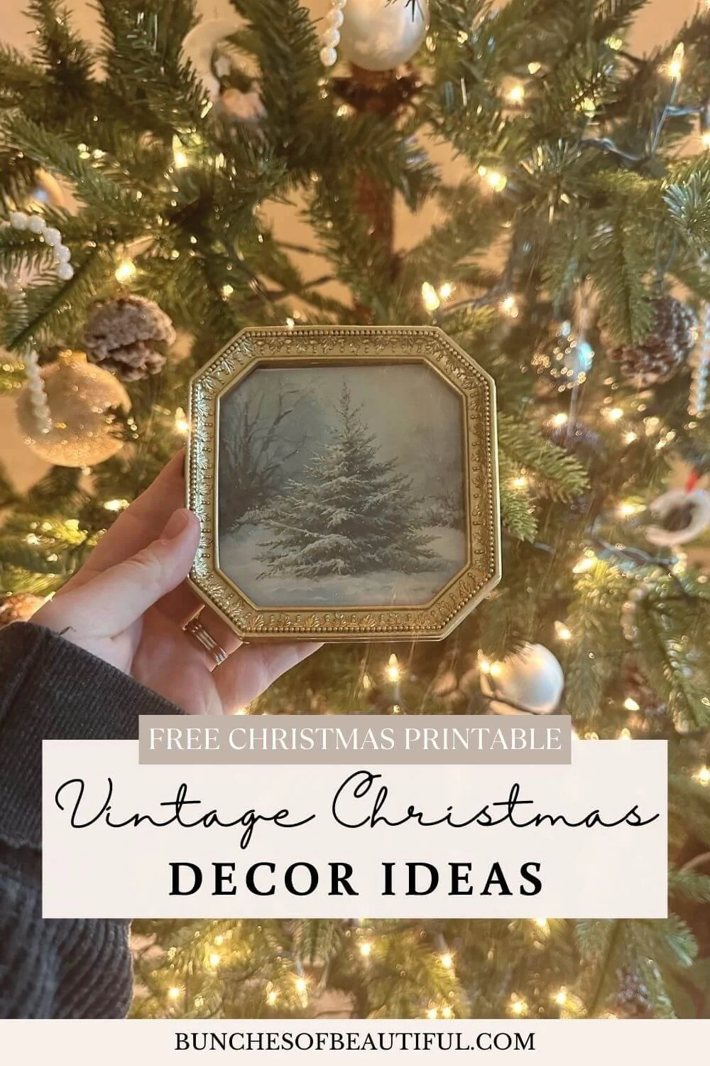 Hand holding a small vintage style gold frame with a snowy Christmas tree illustration in front of a softly lit Christmas tree.