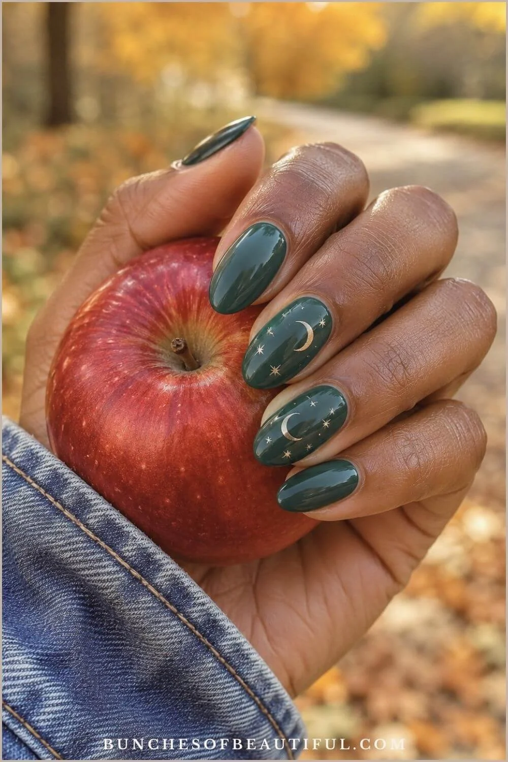 Glossy forest green nails with gold crescent moons and stars while holding a red apple outdoors in fall.