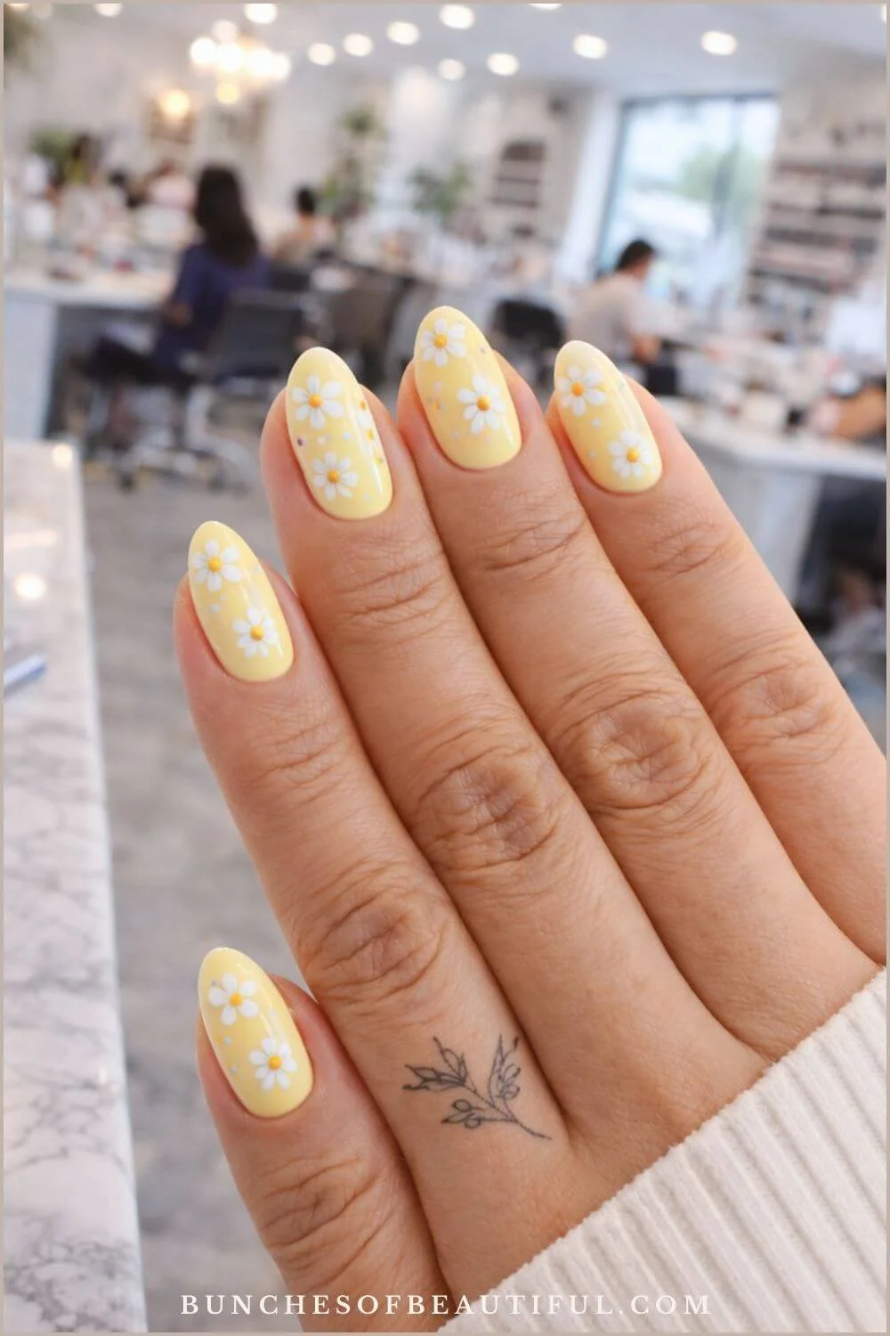 Butter yellow spring nails with white daisy nail art on almond shaped nails in a bright nail salon setting.