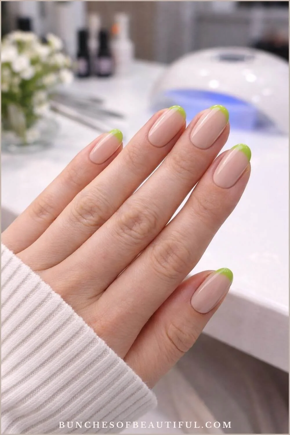 Green French tip spring nails on a nude base photographed in a bright nail salon with a gel nail lamp in the background.