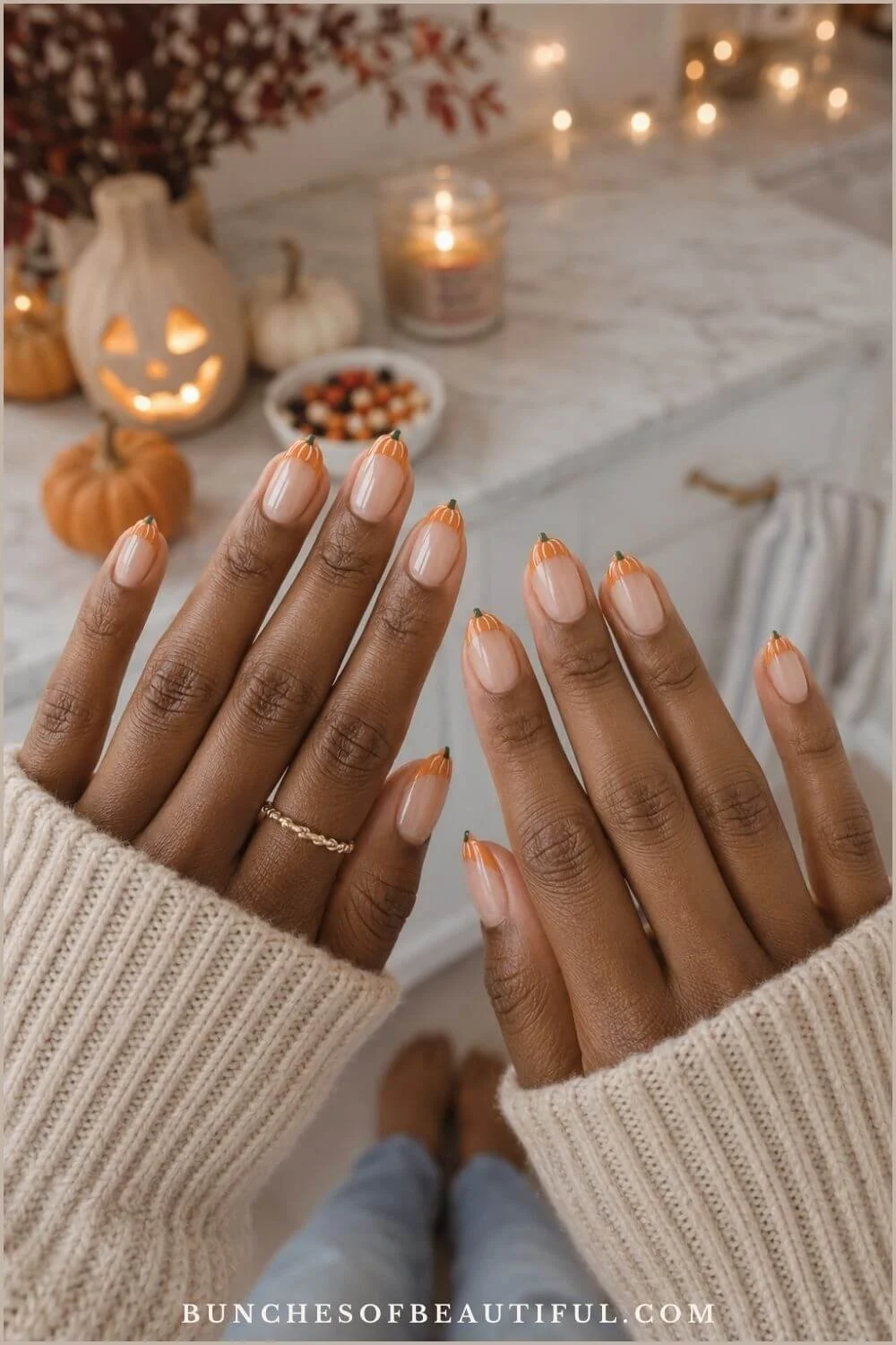 Nude almond nails with tiny orange pumpkin French tips displayed over a cozy Halloween table setting.