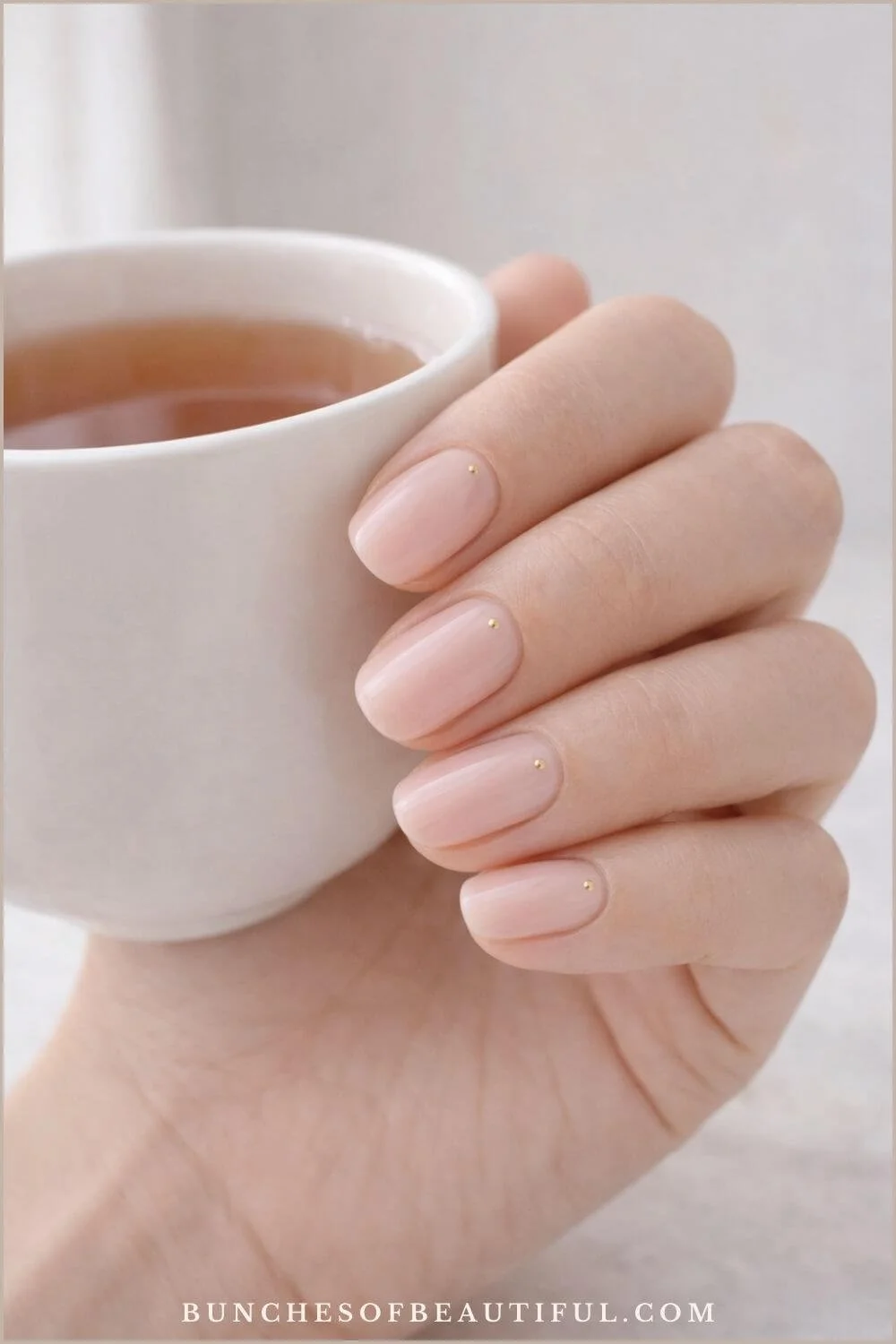Simple spring nails with milky pink polish and tiny gold dot accents on short rounded nails while holding a white coffee mug.