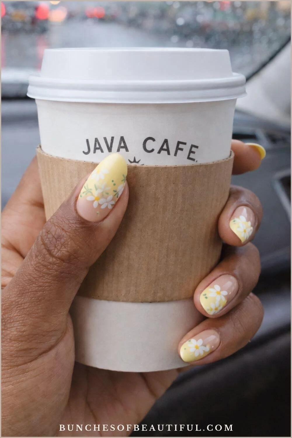Sheer nude spring nails with butter yellow floral French tips, photographed holding a coffee cup in a car on a rainy day.