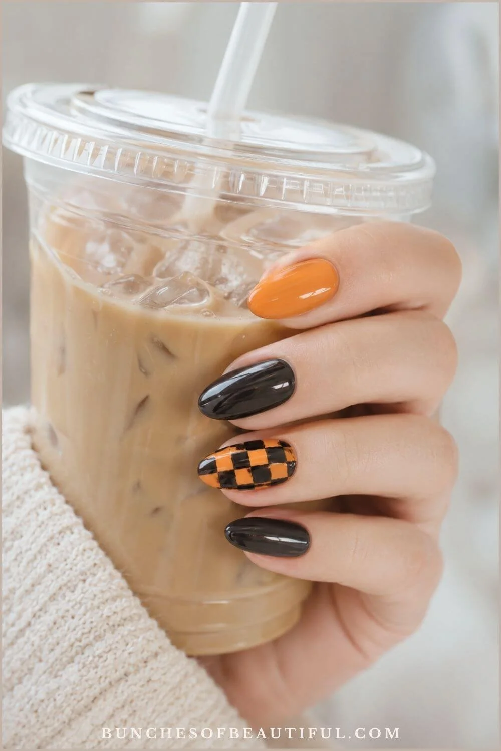 Orange and glossy black oval nails with a hand-painted checkerboard accent while holding an iced coffee.