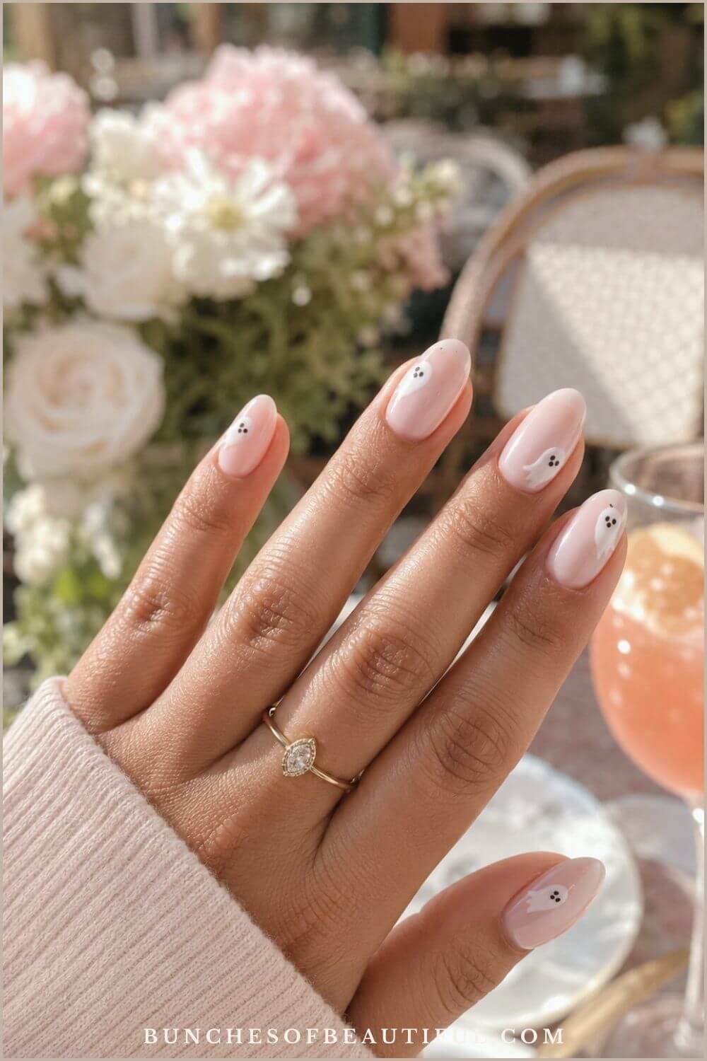 Sheer pink almond nails decorated with small white peekaboo ghosts at an outdoor café table.