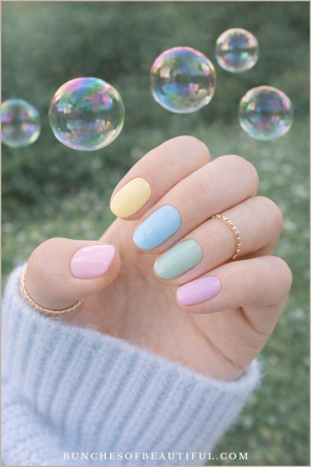 Short pastel rainbow spring nails with each nail a different pastel color including yellow, blue, green, pink, and purple photographed outdoors with bubbles in the background.