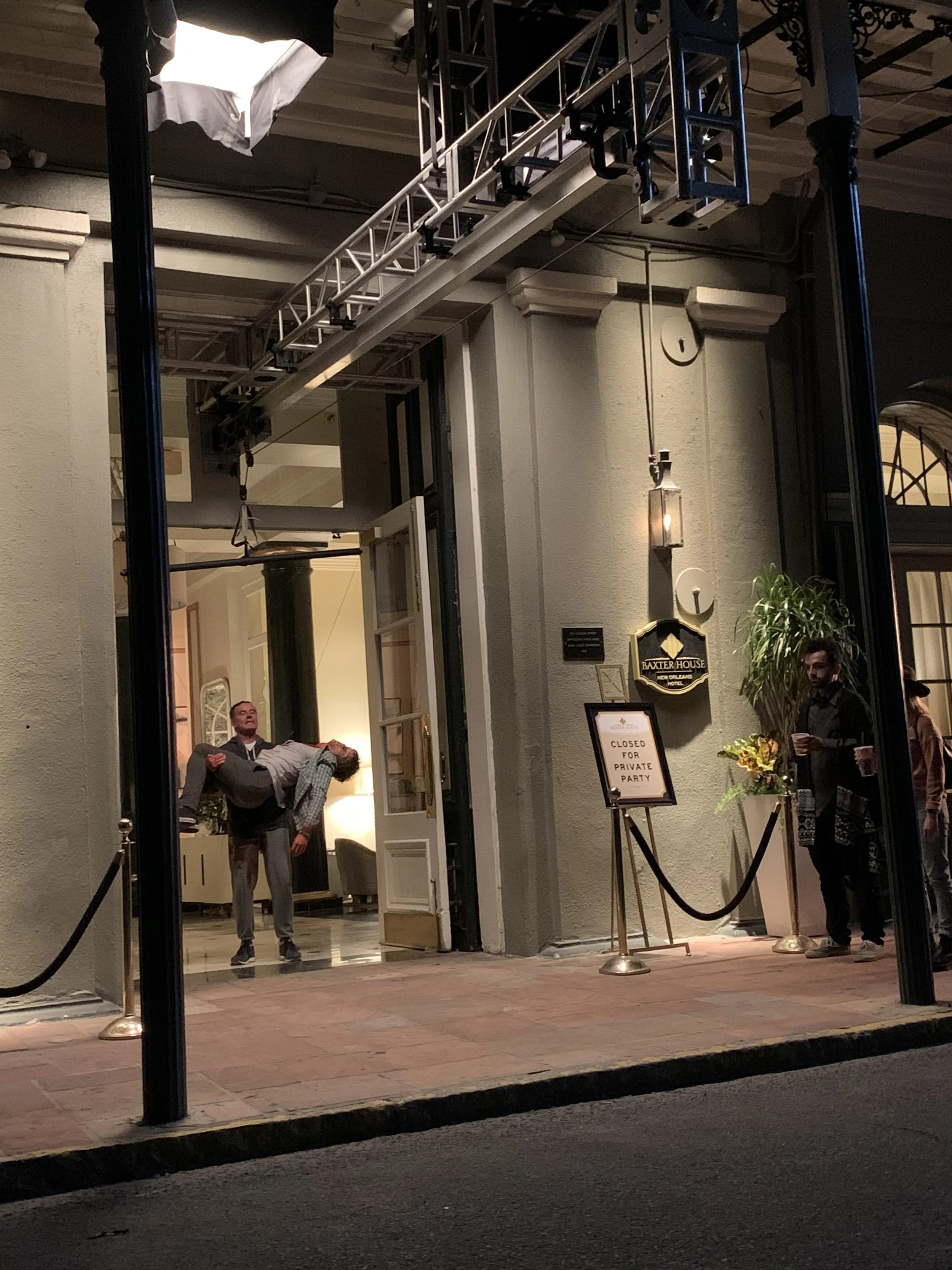 People standing outside a building with a sign indicating it is closed for a private party. Someone is holding a man who is dipping him in a dance pose, and other people are near the entrance.