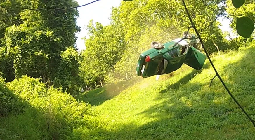 A green car has flipped upside down on a grassy hill in a wooded area.