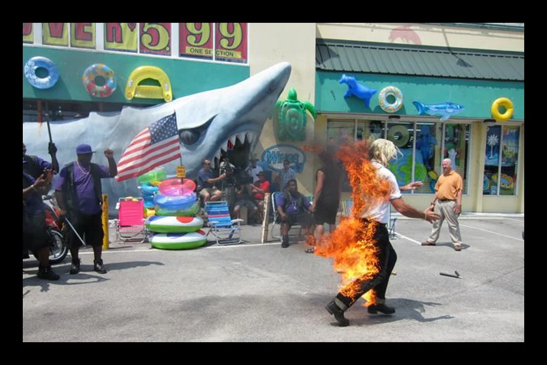 A man with blonde hair is on fire outside an amusement park or water park with a large shark sculpture and inflatable pool toys in the background.