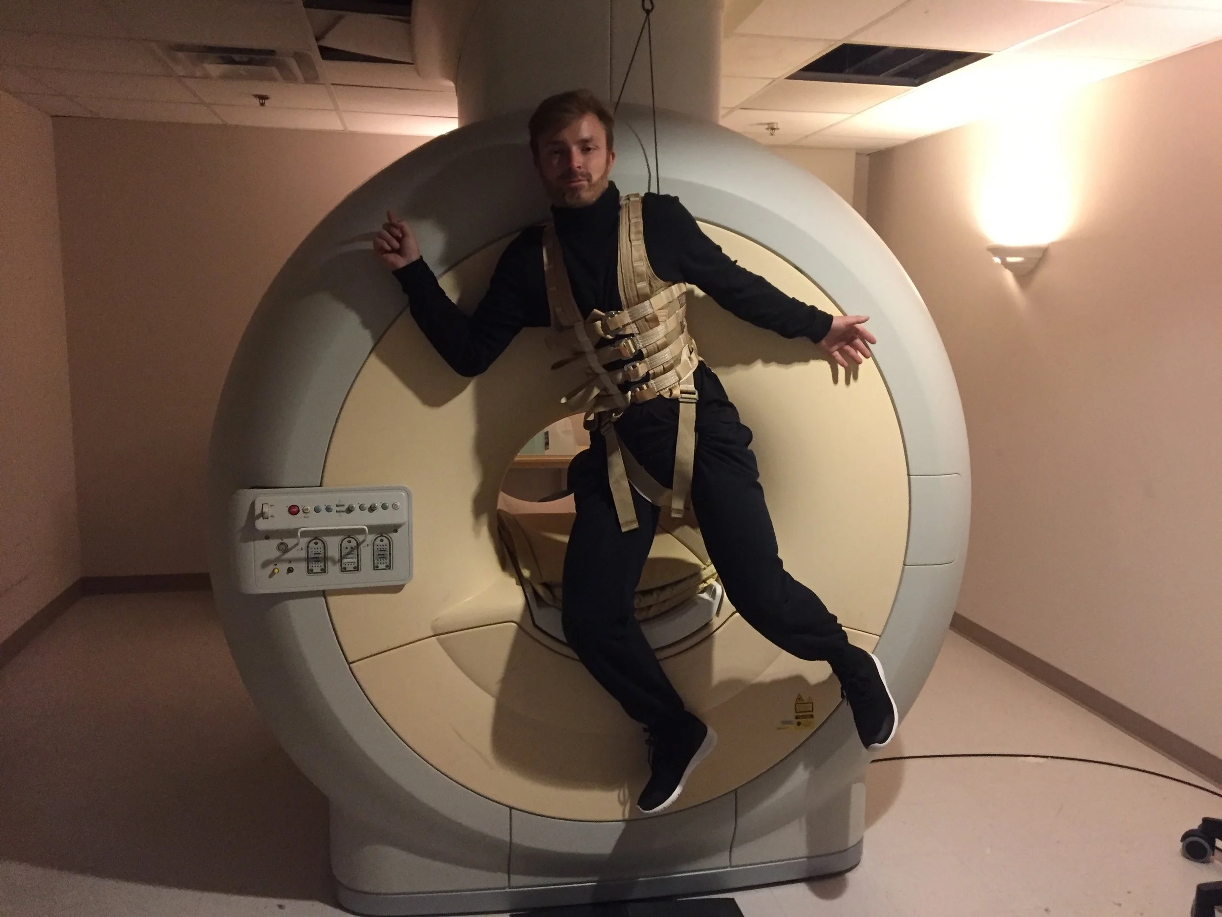 A man is lying inside an MRI machine with his arms and legs outstretched, dressed in black clothing and a beige harness for support.