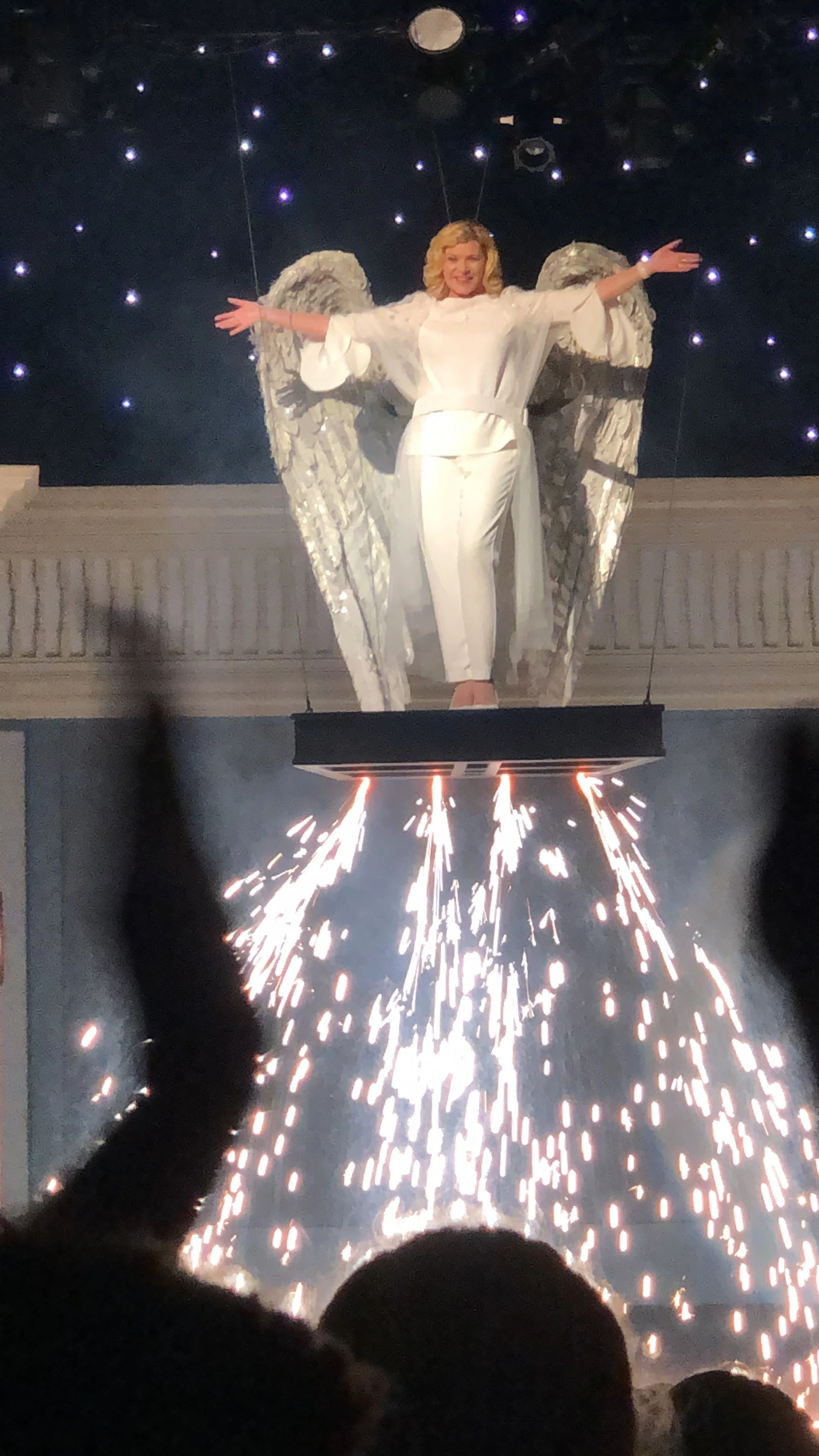 A woman dressed in white standing on a platform with large angel wings, arms outstretched, with fireworks or sparks falling below her, in front of a background with small bright stars.