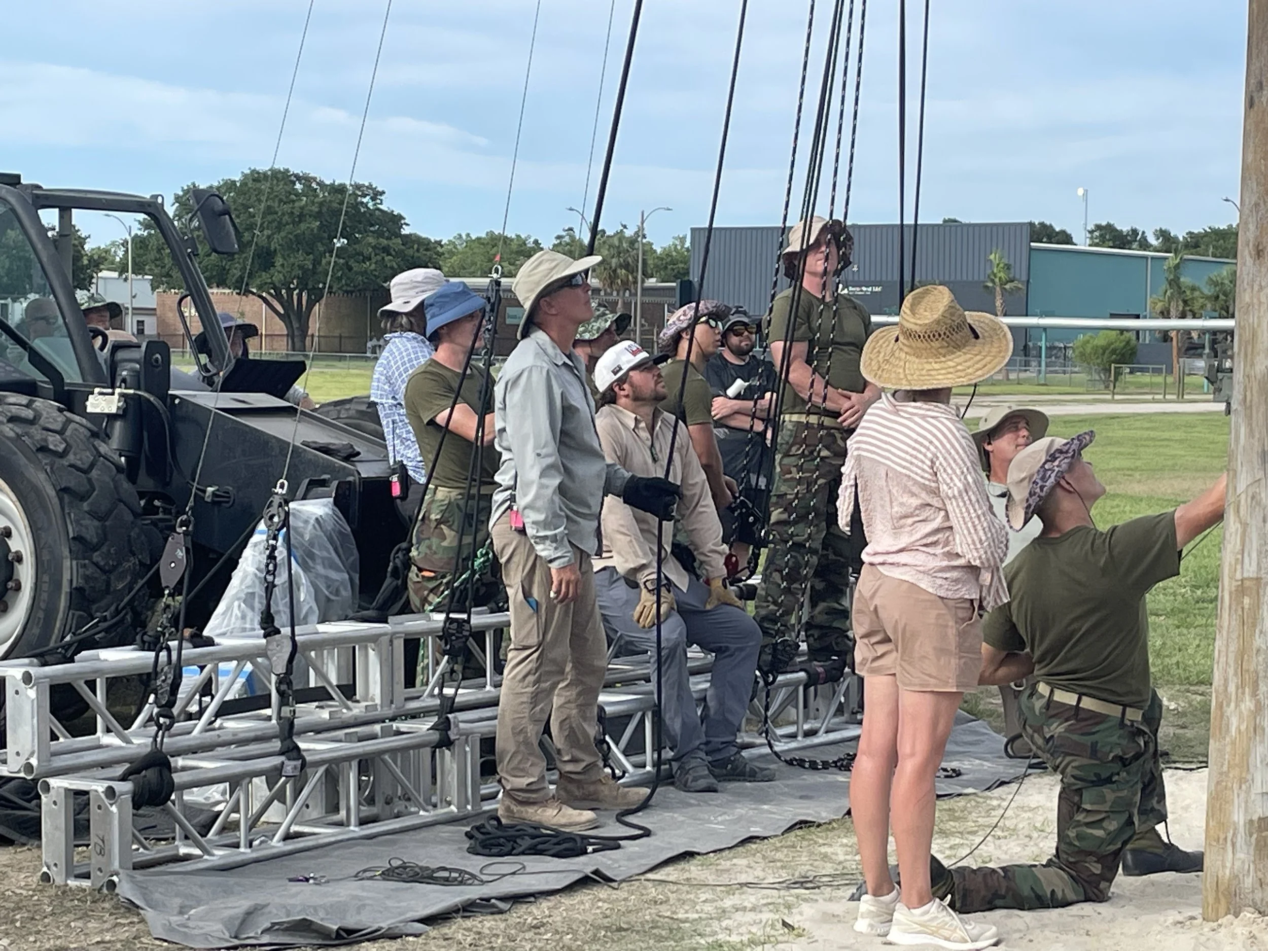 A group of people on a construction platform, reaching upward, with some looking through binoculars or glasses, set outdoors during daytime.