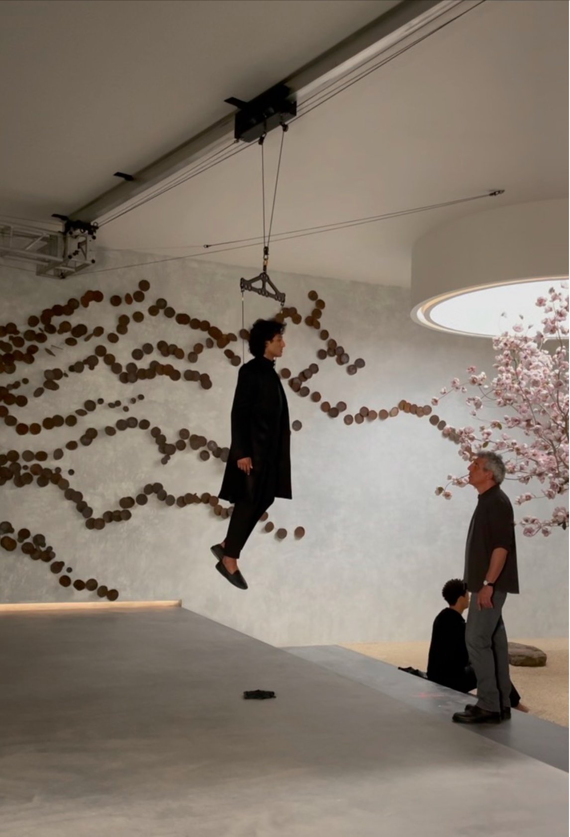 A man in a black coat appears to be levitating or suspended in the air inside a modern, minimalist space with decorative wall art and pink blossoms on a tree.