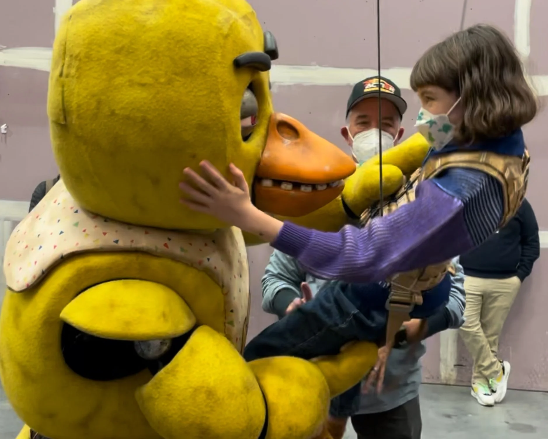 A person in a costume with the head of a yellow cartoon character, holding hands with a young girl who is touching the character's face. Both are wearing face masks. Two other people are in the background, also wearing masks, in an indoor setting with a pink wall.