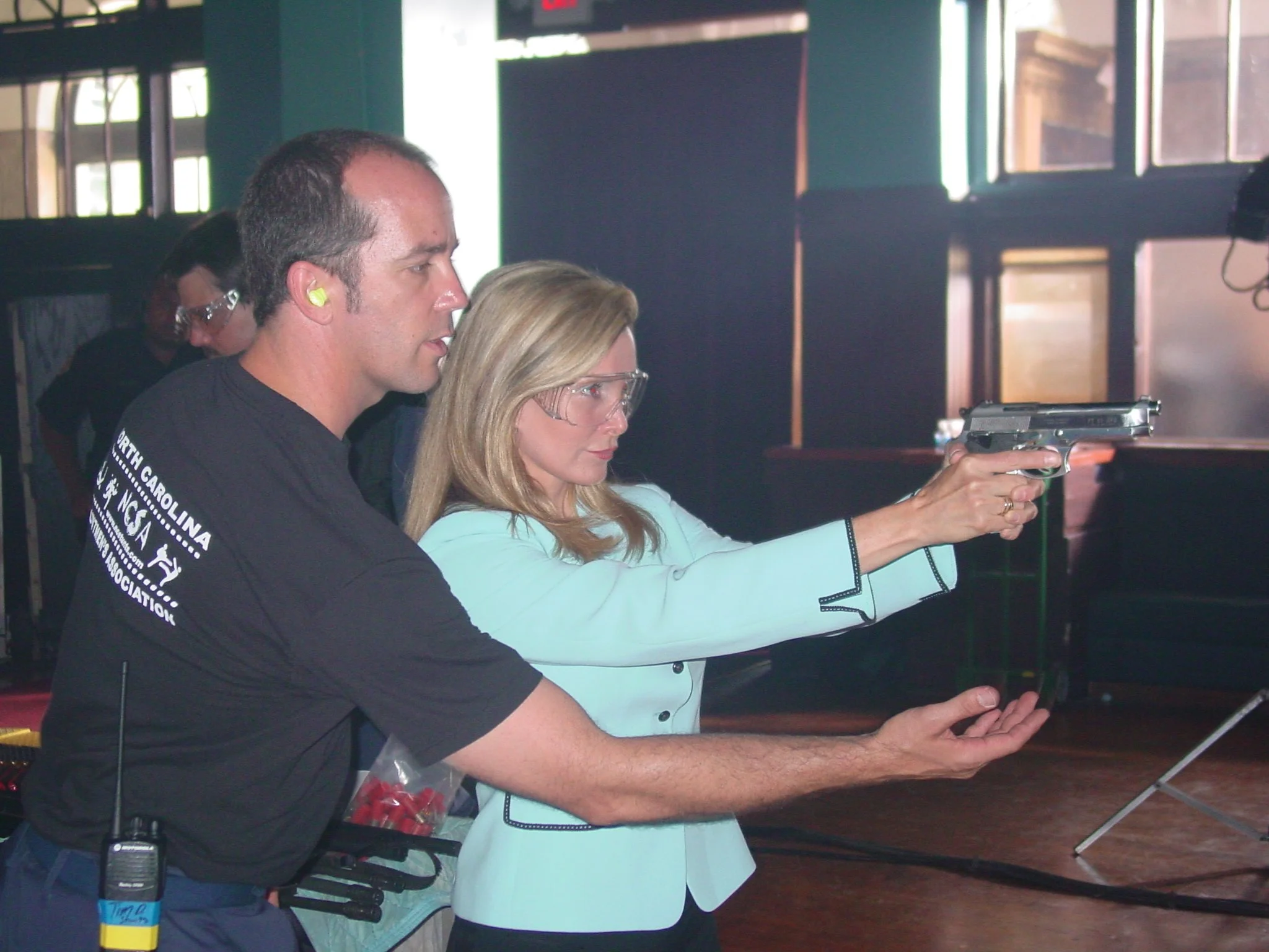 A woman in a light blue blazer is aiming a handgun, guided by a man in a black shirt at an indoor shooting range.