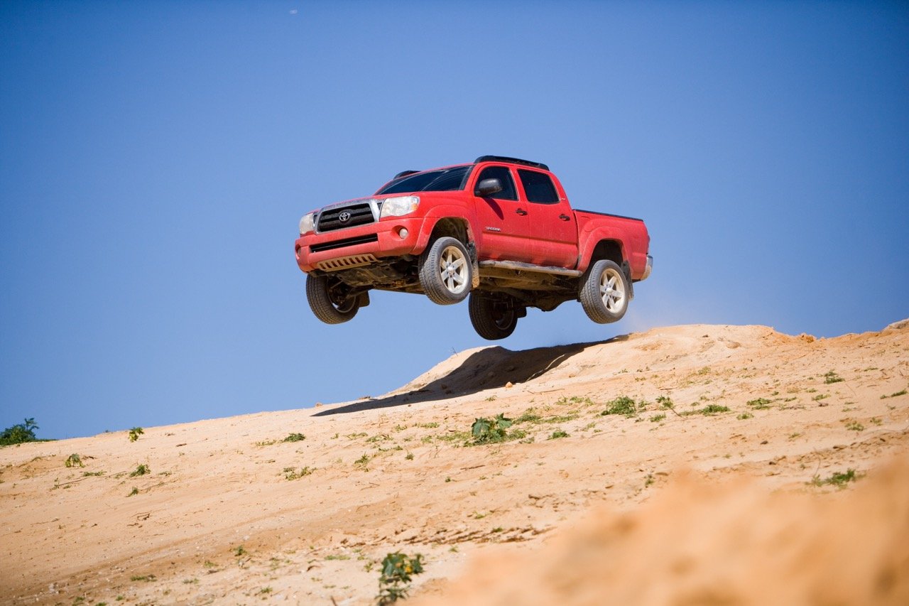 A red pickup truck in mid-air during a jump on a dirt terrain, with a clear blue sky in the background.