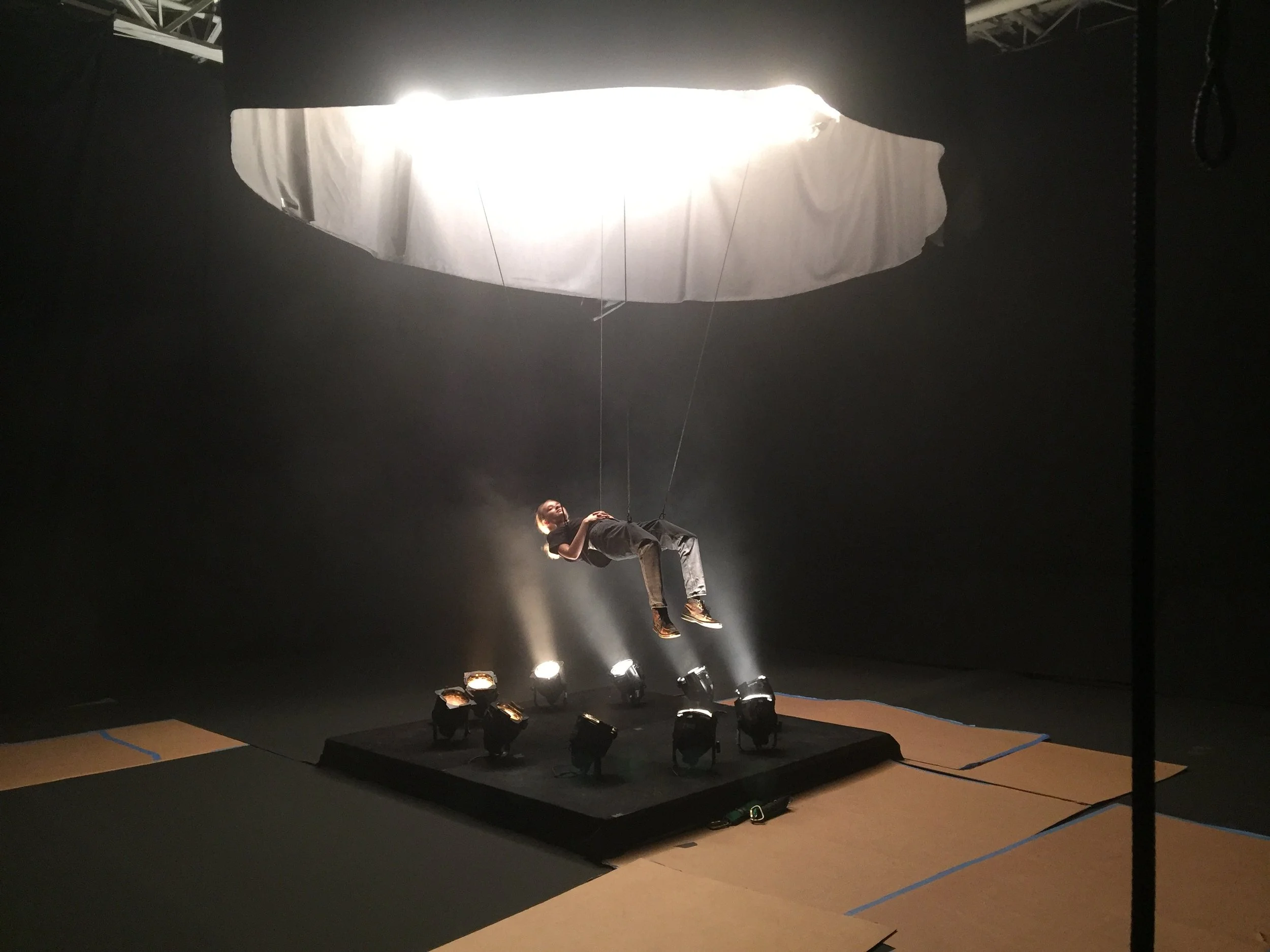 Person lying on a harness under a large white fabric circle suspended from the ceiling, with multiple spotlights pointed towards the person. The scene appears to be in a dark room, possibly a stage or studio setup.