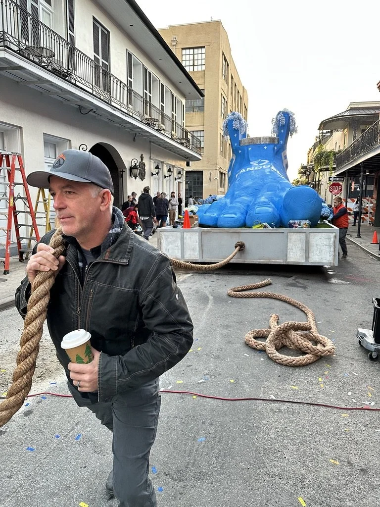 A man holding a coffee cup and a thick rope on a city street with a large float of a blue Land Rover Defender in the background, surrounded by people and construction cones, during a parade or festival.