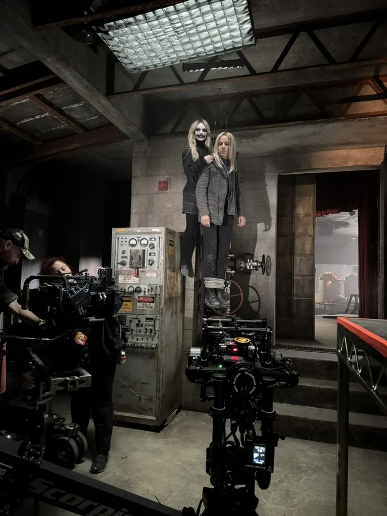 Two women, one with ghostly makeup, standing on a platform with the other tied up with ropes in an industrial setting, possibly behind the scenes of a film or TV production.
