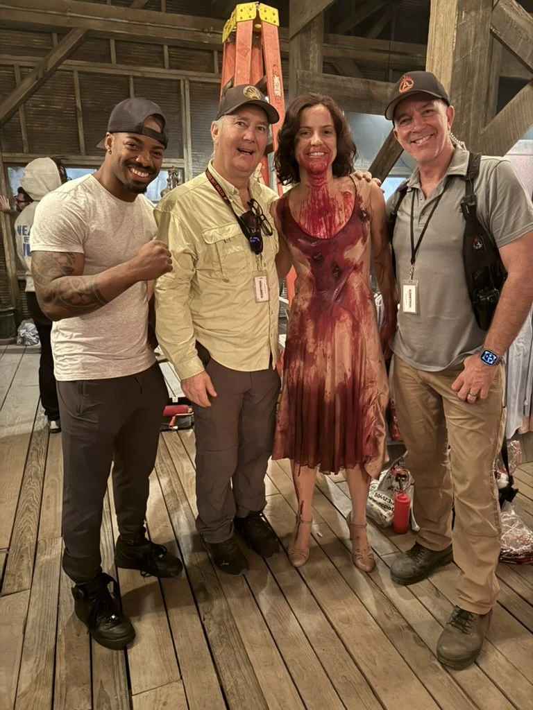 Four people standing together inside a wooden barn, with a ladder behind them, celebrating a Halloween or horror-themed event. The woman in the center is dressed as a zombie or a haunted figure, with fake blood on her face and body, wearing a blood-stained dress. The three men are smiling and dressed casually.