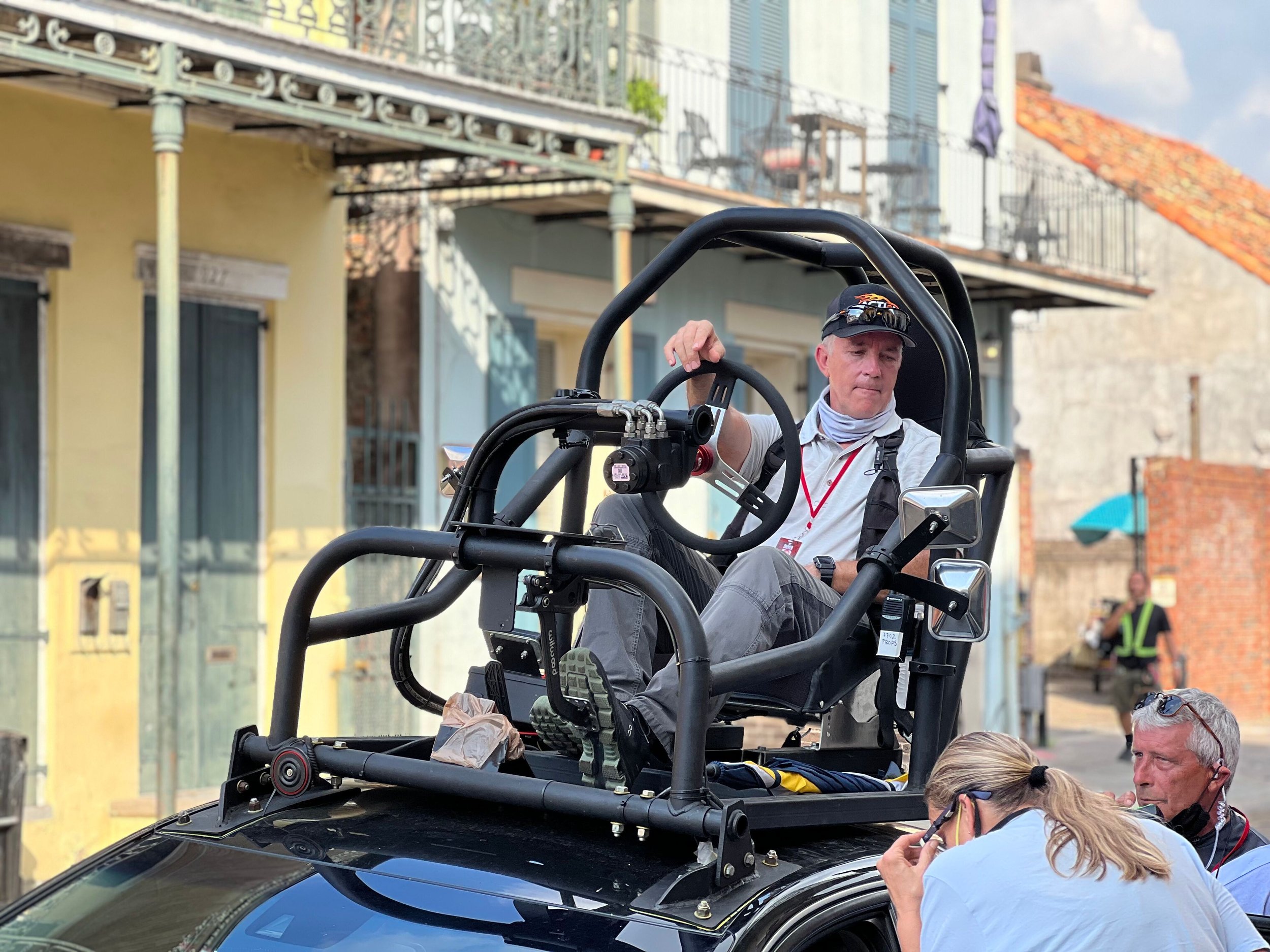 A man sitting in a racing-style seat on a vehicle with a roll cage, surrounded by crew members on a street with colorful buildings and other people.