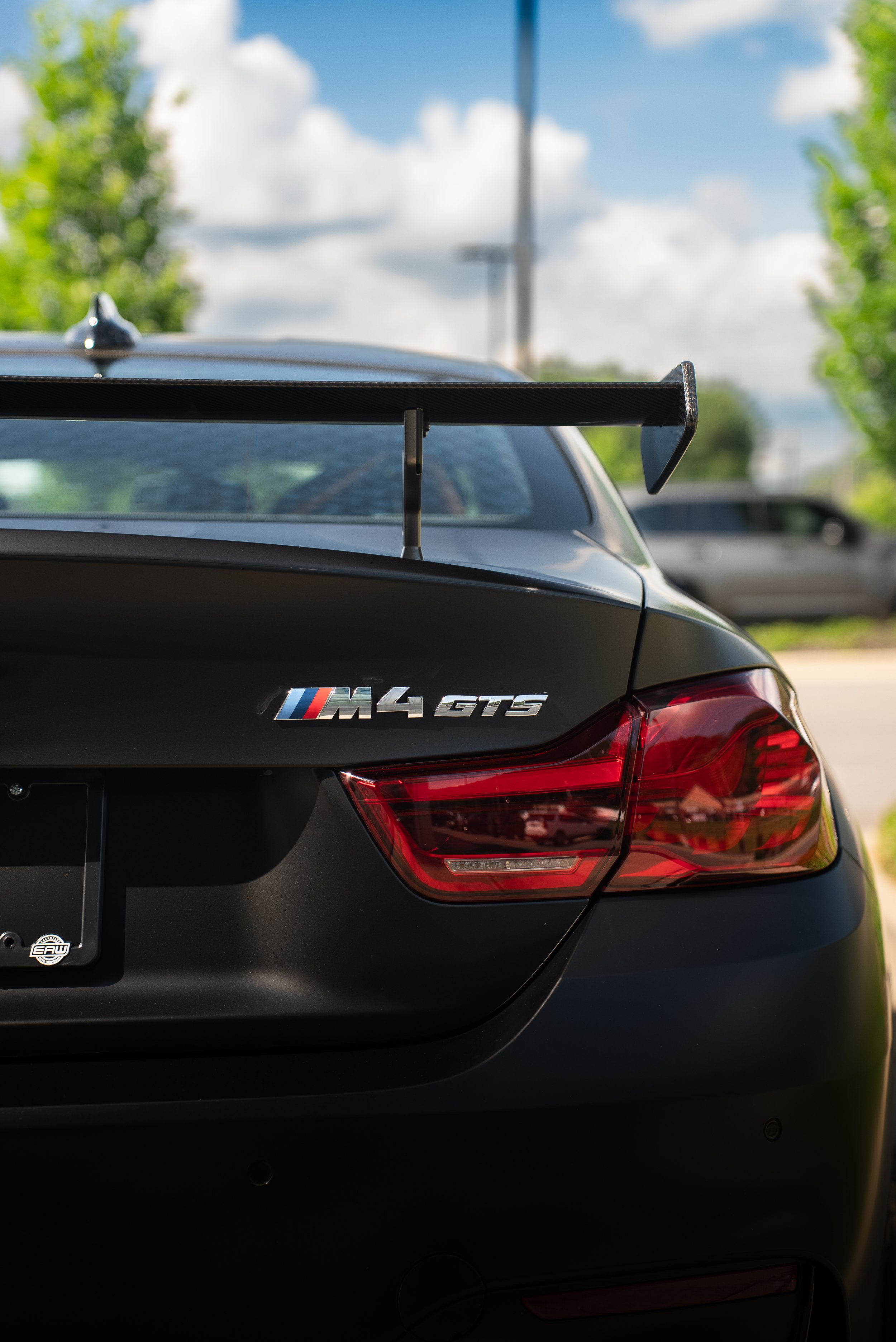 The rear of a black BMW M4 GTS sports car with a large rear wing, parked outdoors with other vehicles and greenery in the background.