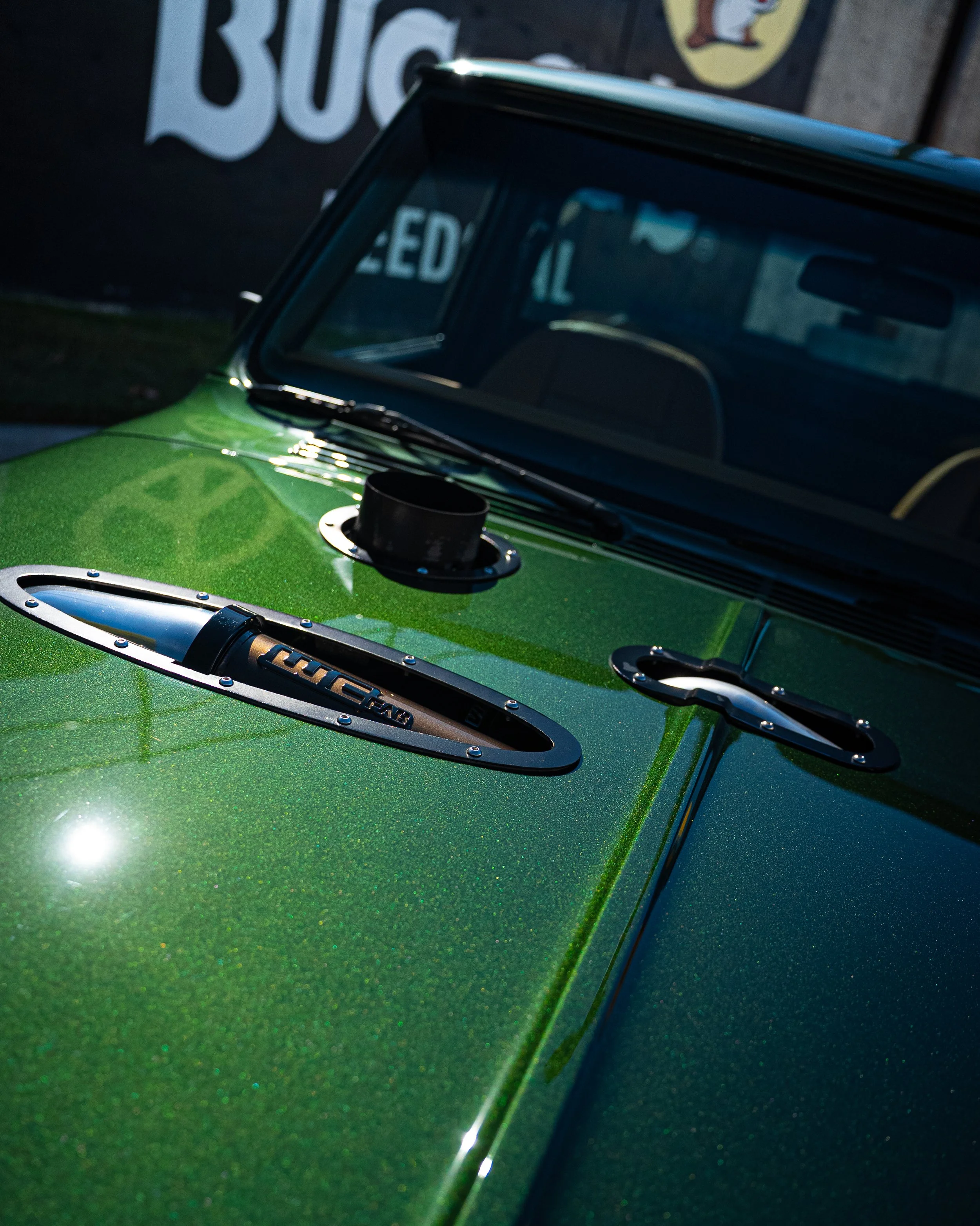 Close-up of a vintage green car with a glittery finish, showing a part of the windshield and the hood with metallic accents and ventilation openings.