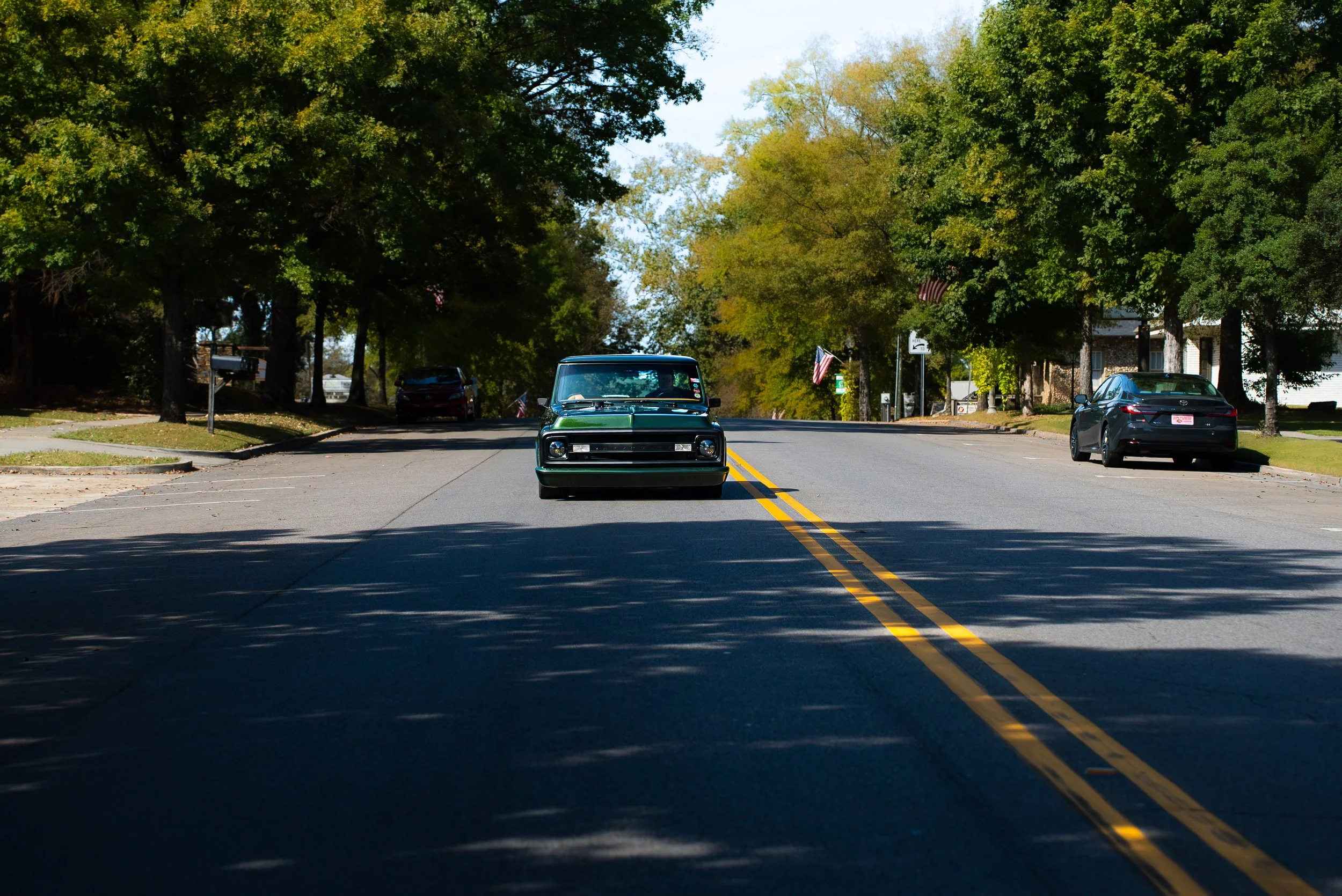 A small vintage black car driving down a suburban street lined with green trees and parked cars, under a partly cloudy sky.