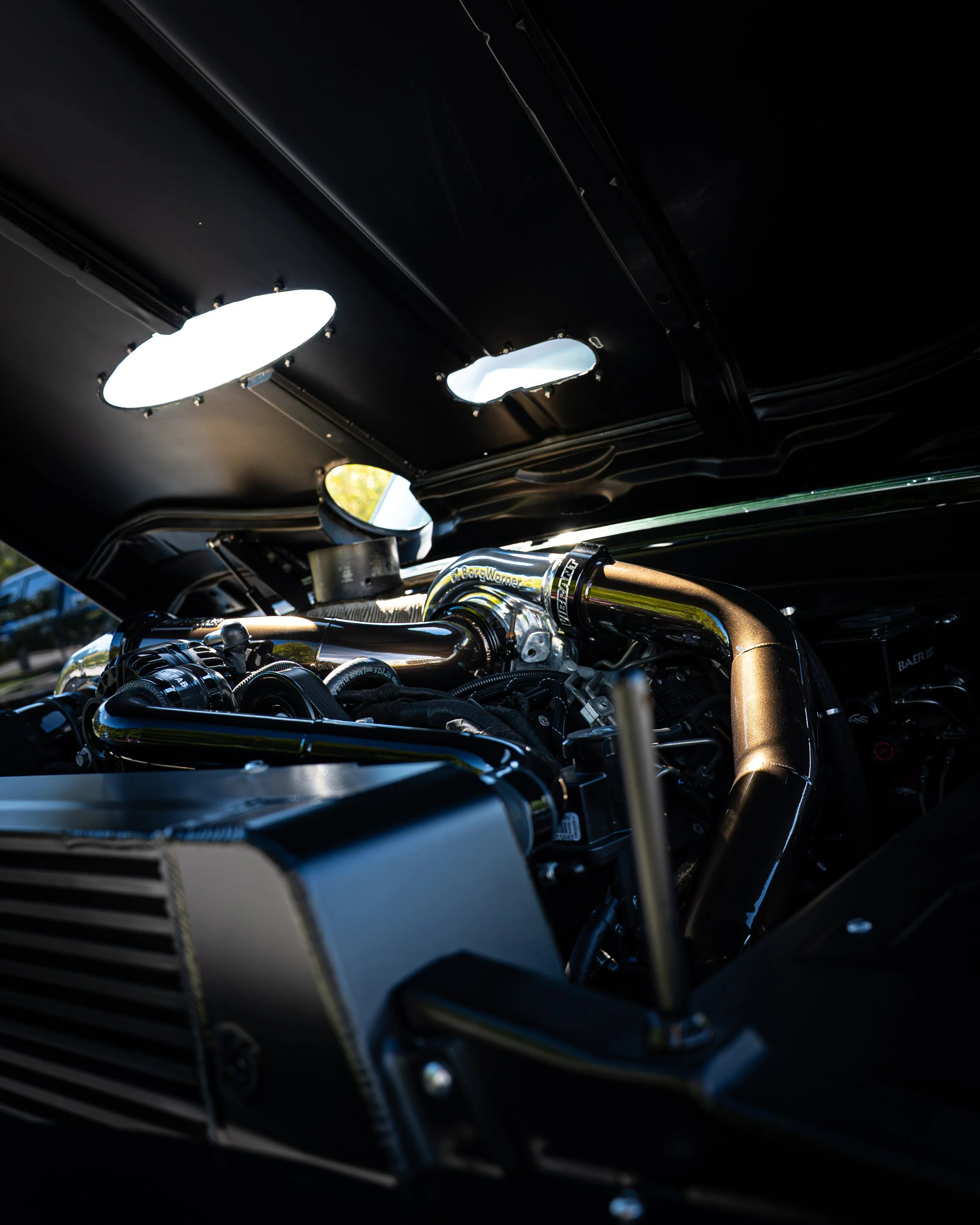 Close-up photo of a motorcycle engine under a black hood with two illuminated oval and irregular holes in the hood, and some shiny chrome and metallic parts of the engine visible.