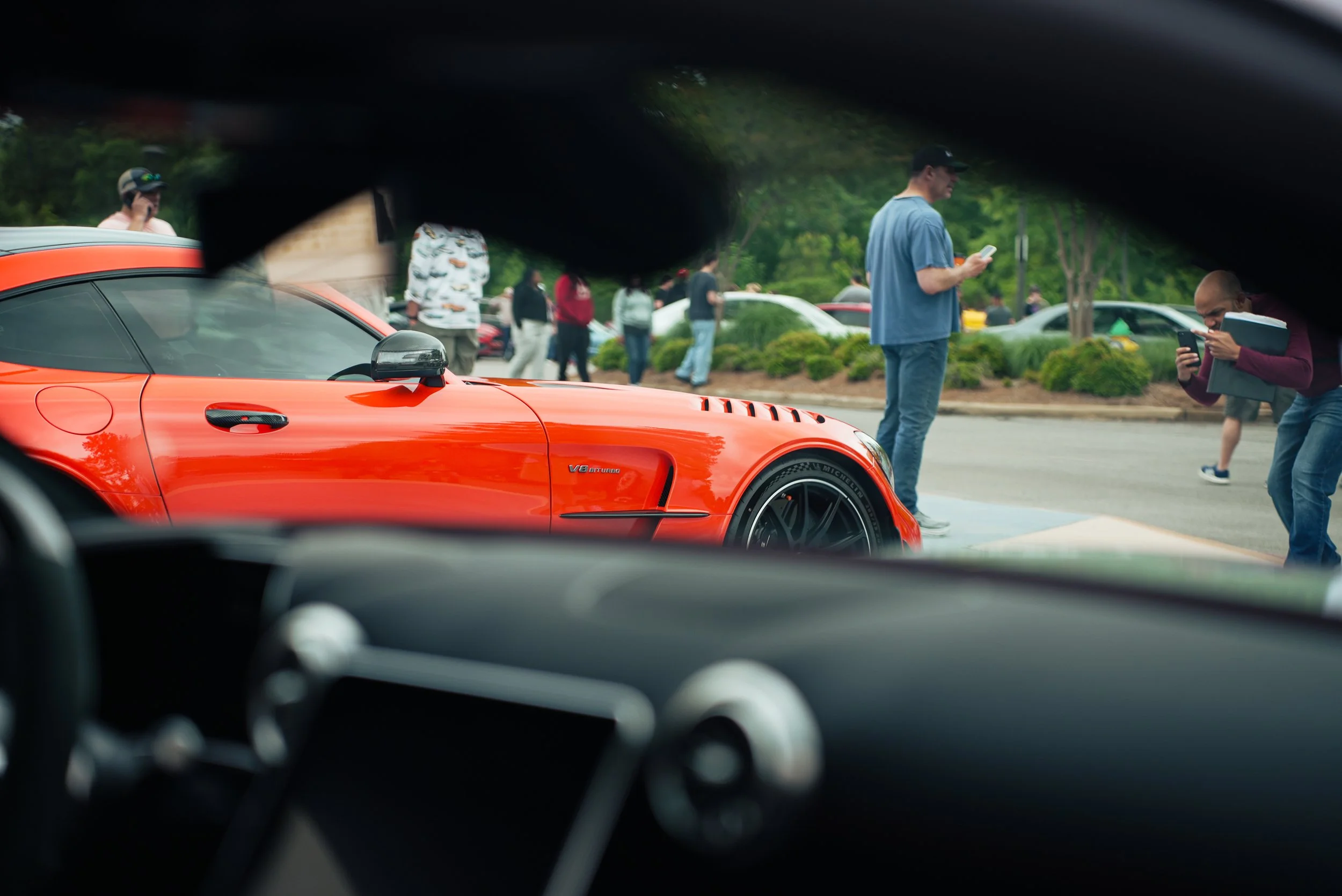 A view from inside a car looking out through the windshield at a parking lot with people and cars, with a bright orange sports car in the foreground.