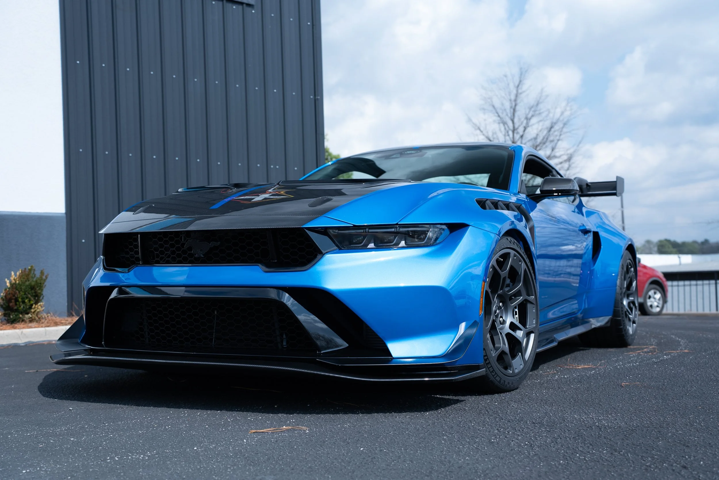 A blue sports car with black accents, parked outdoors near a modern building with a dark gray wall, under a partly cloudy sky.