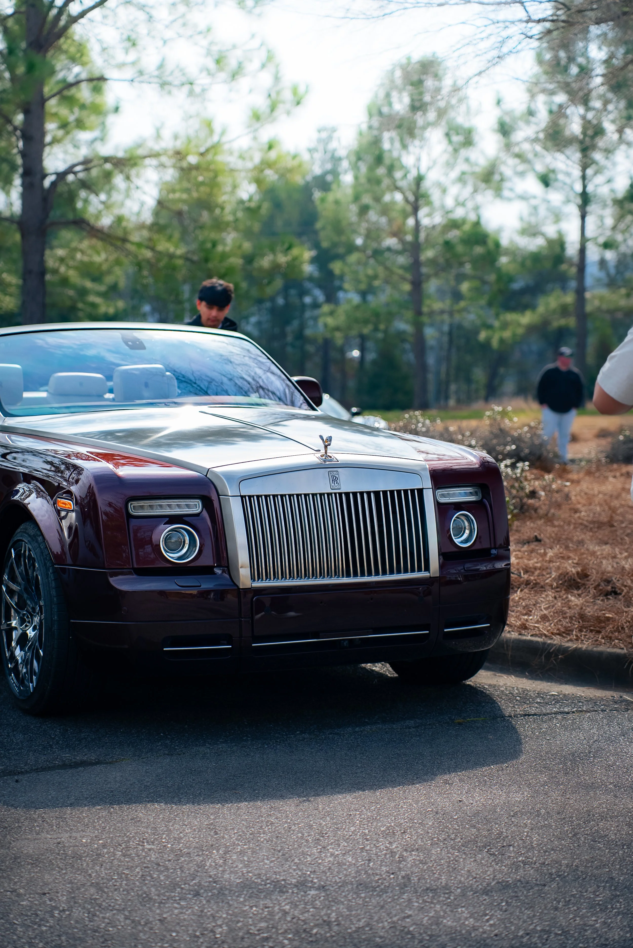 A burgundy and silver luxury Rolls-Royce car parked on a paved street, with a man in a dark jacket looking at it and two other men standing in the background among trees.