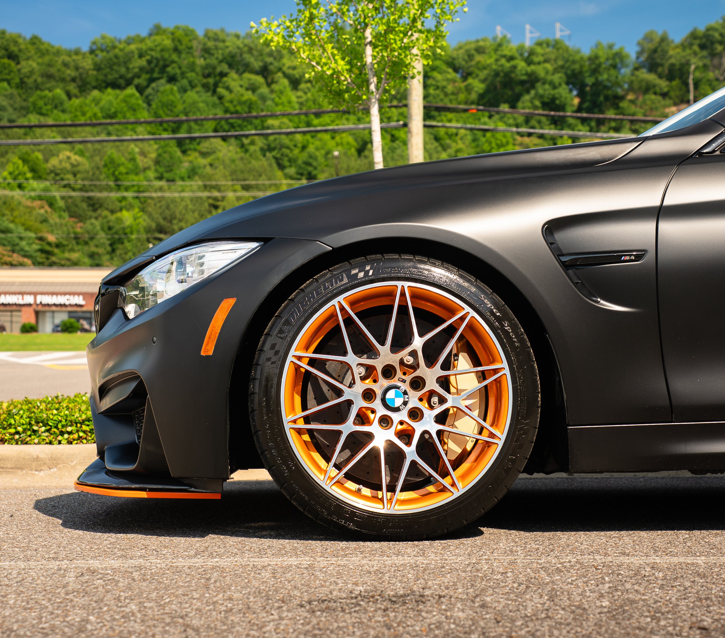 Close-up of a black sports car with orange wheels parked on asphalt, with green trees and a clear blue sky in the background.