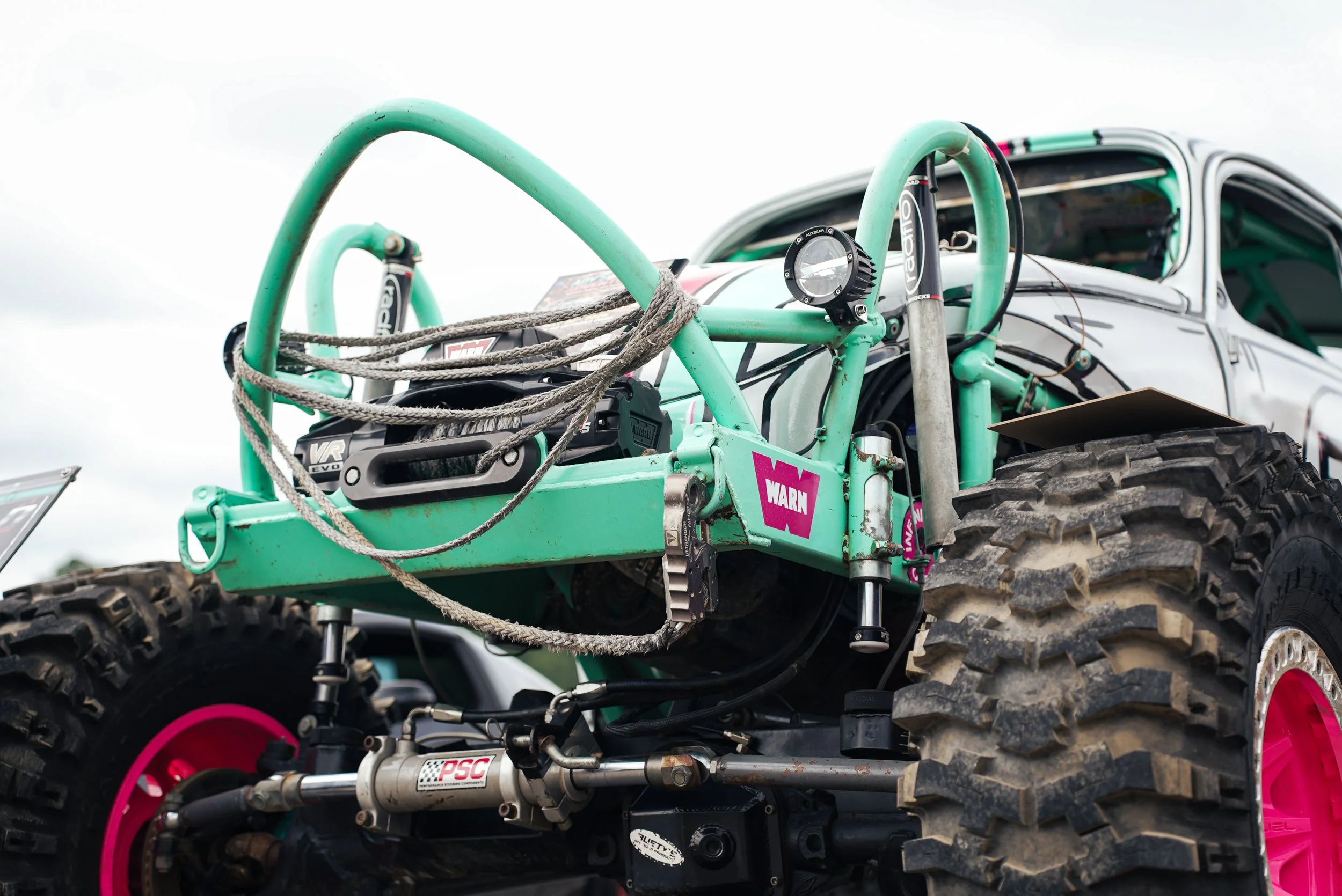 Close-up of off-road racing vehicle with green roll cage, large tires with pink rims, and various mechanical parts, parked outdoors under overcast sky.