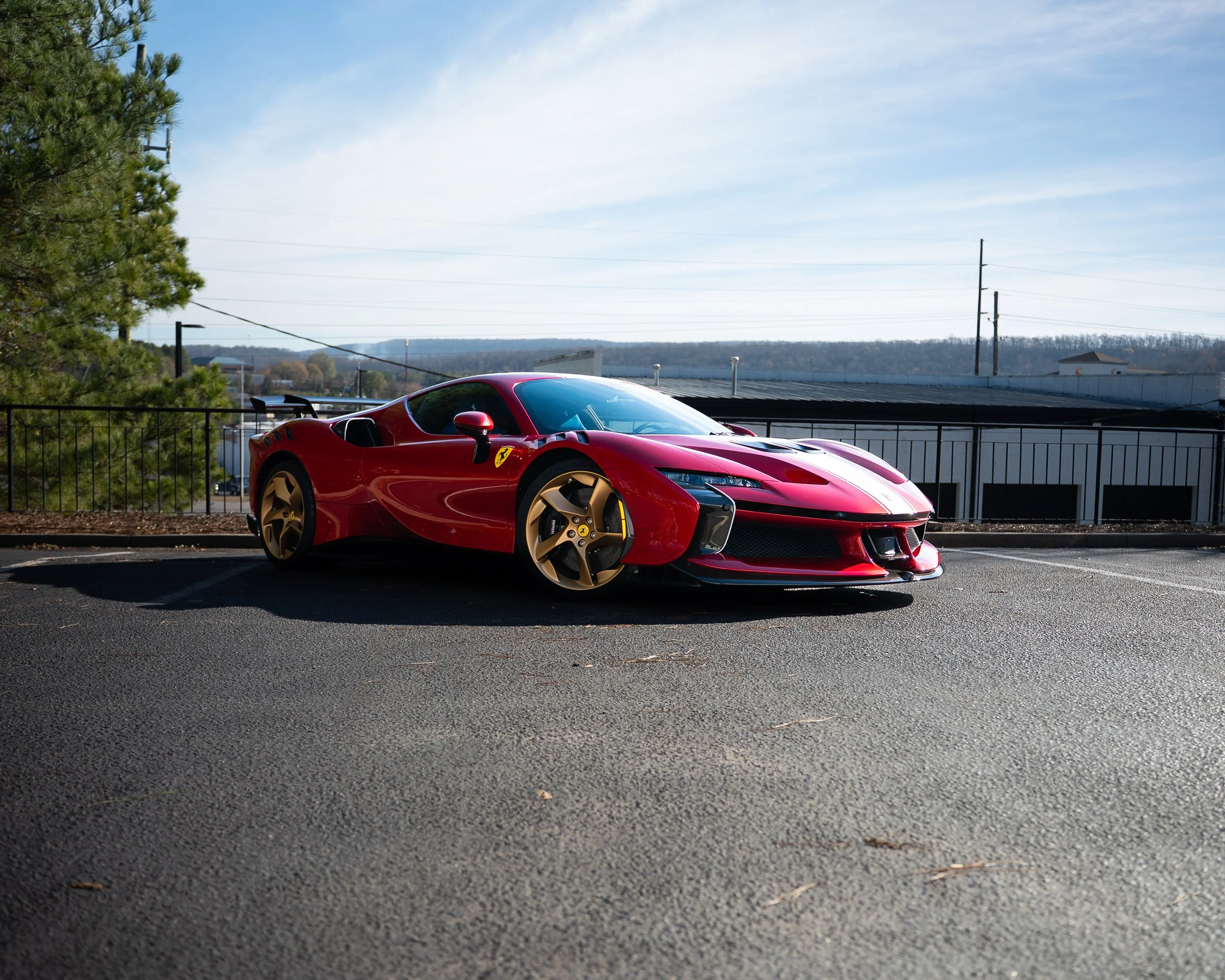 Red Ferrari sports car parked outdoors with a fence, trees, power lines, and a building in the background under a partly cloudy sky.