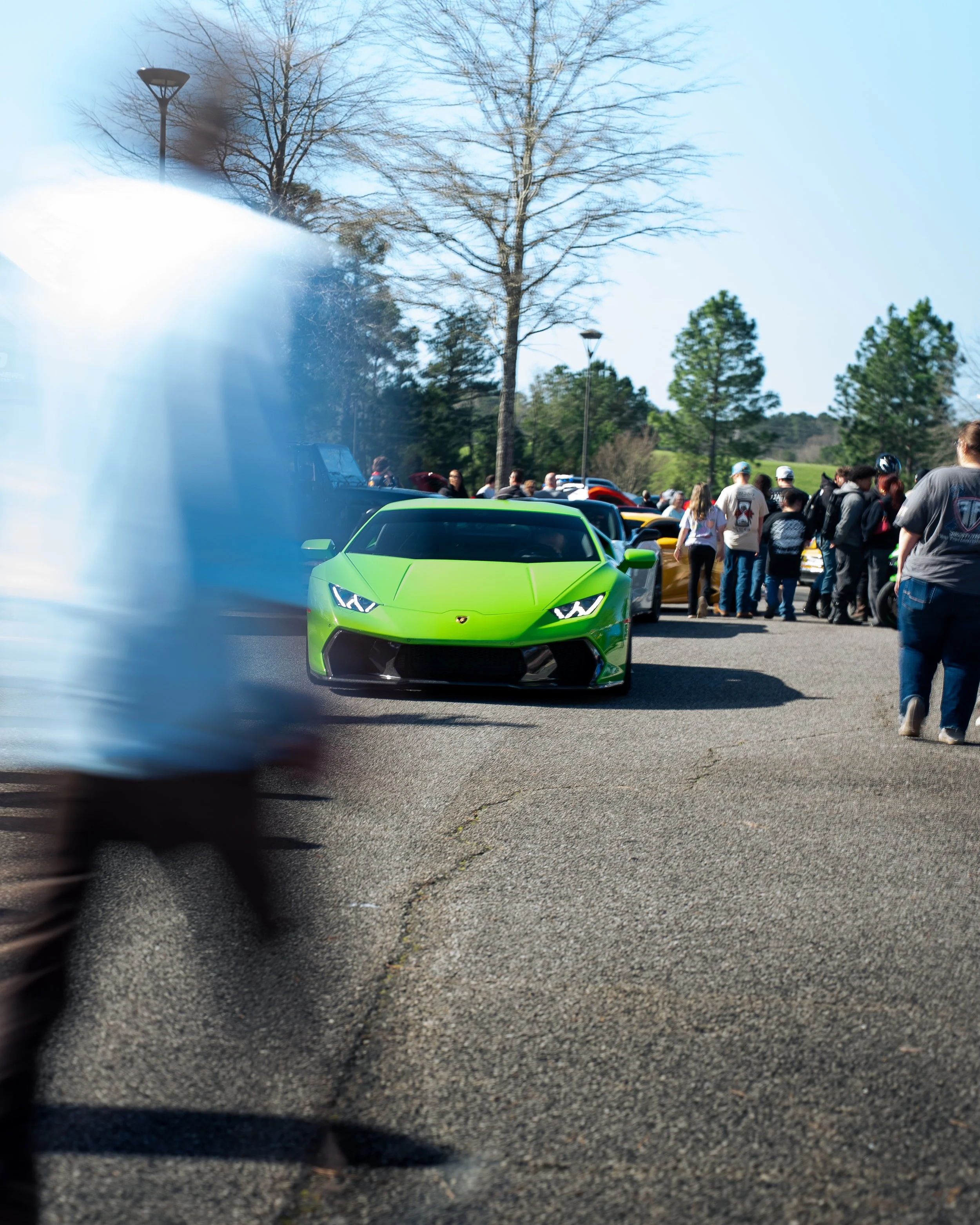 A group of people gathered outdoors near a parking lot with a bright green Lamborghini sports car in the foreground, with trees and a lamppost in the background, and a motorcyclist's blurred wheel visible at the left edge of the image.